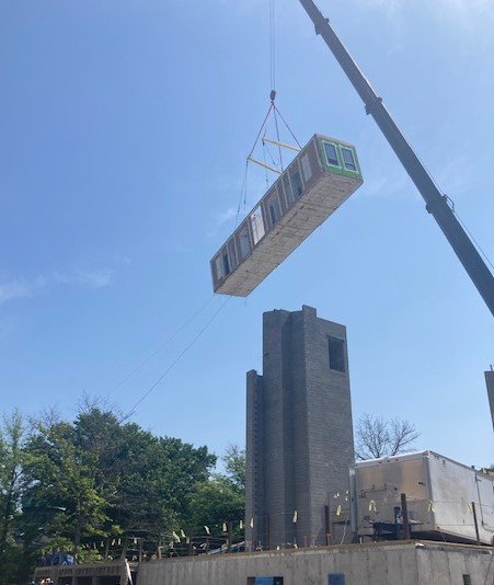 A crane lifting a large modular building into a construction site with a tall brick chimney in the background.