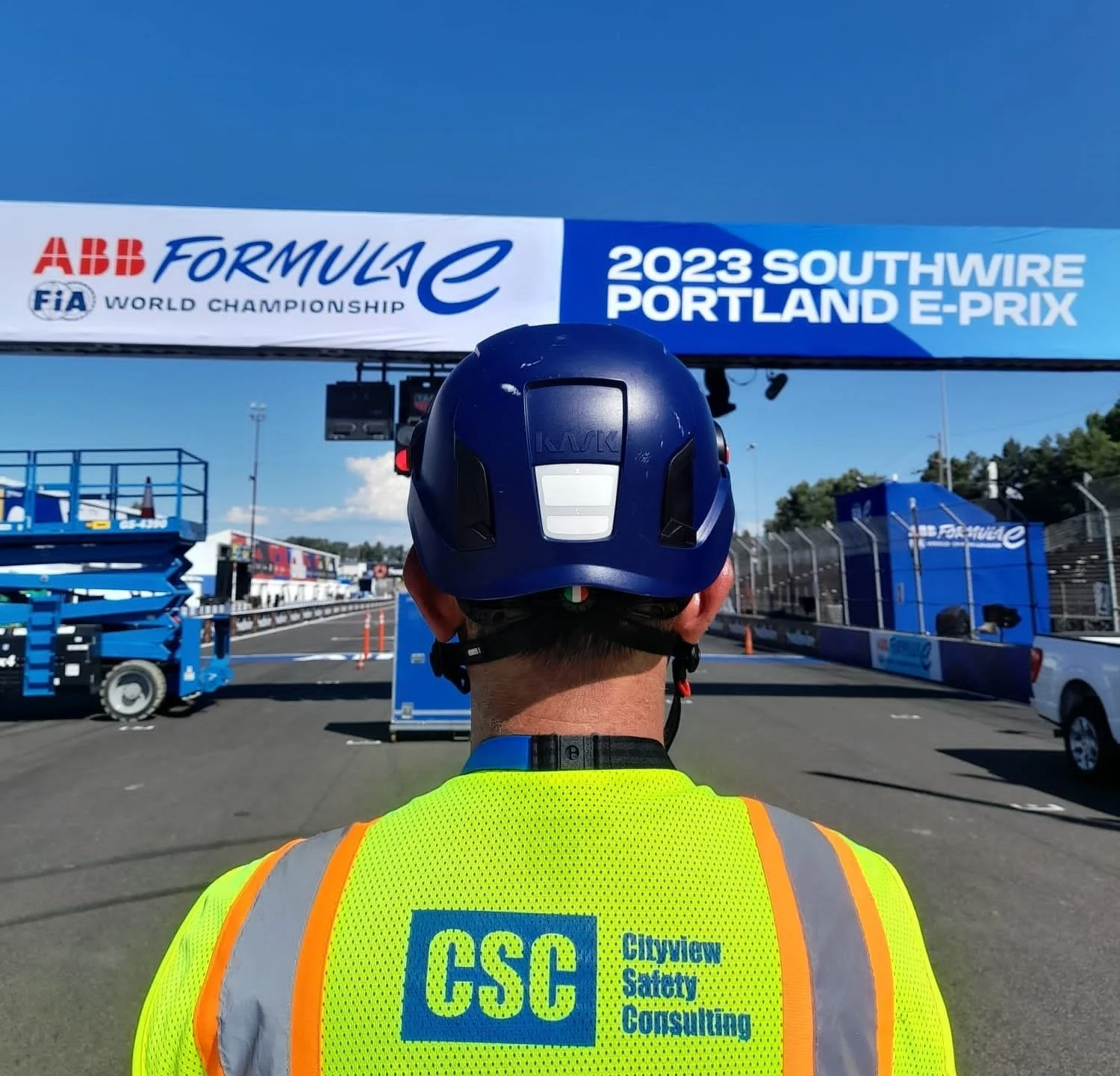 A person wearing a blue helmet and a bright yellow safety vest with 'CSC Cityview Safety Consulting' on the back is standing in front of the start/finish line at the 2023 Portland Southwire E-Prix, a Formula E race event.