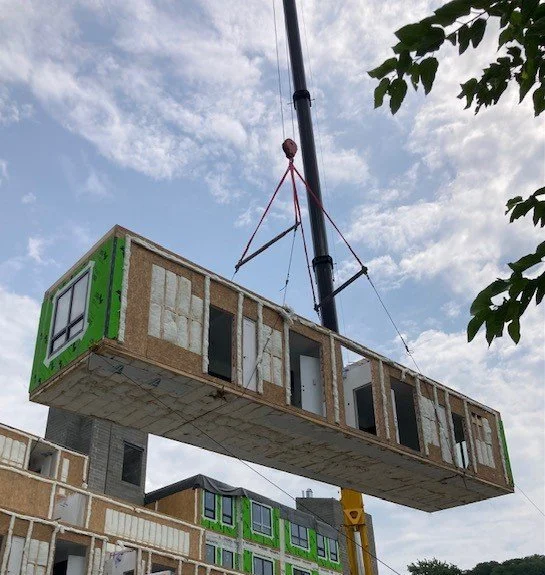 A large construction crane lifting a prefabricated building module at a construction site during the daytime with a partly cloudy sky.
