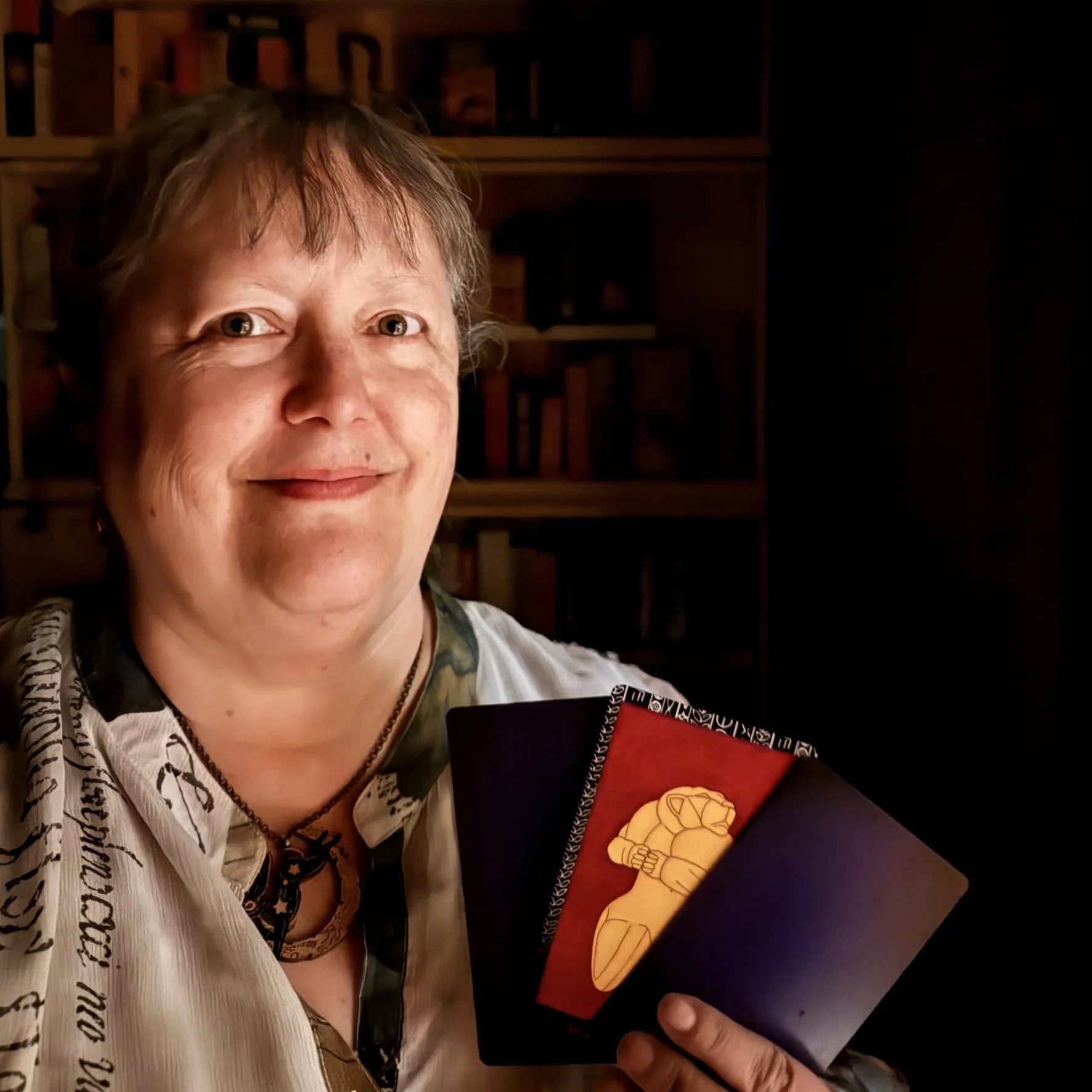 A woman with dark hair holding three tarot cards, with several shelves of tarot decks in the darkness behind her.