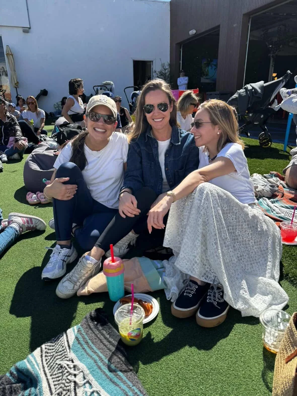 Three women sitting on the grass at an outdoor gathering, smiling, with many people in the background, some sitting and some standing, drinks nearby, sunny weather.