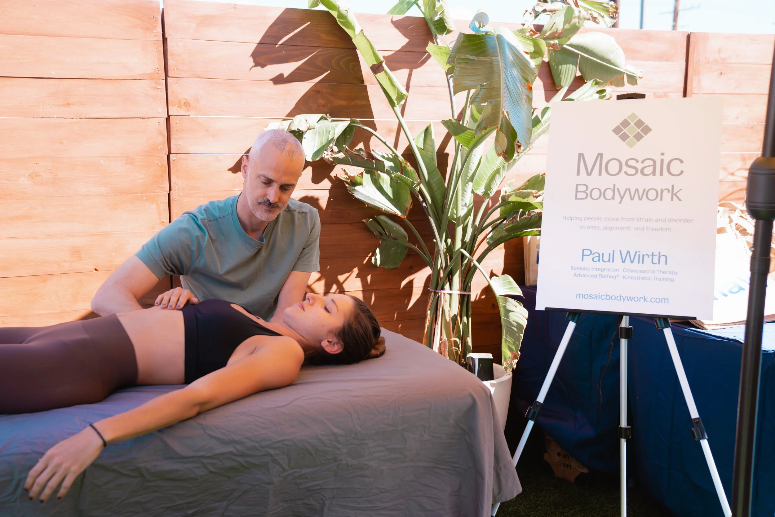 A person receiving a bodywork treatment from a practitioner on a massage table in an outdoor setting with a wooden fence and large green plant nearby. A sign next to them displays the name 'Mosaic Bodywork' and the practitioner's name and credentials