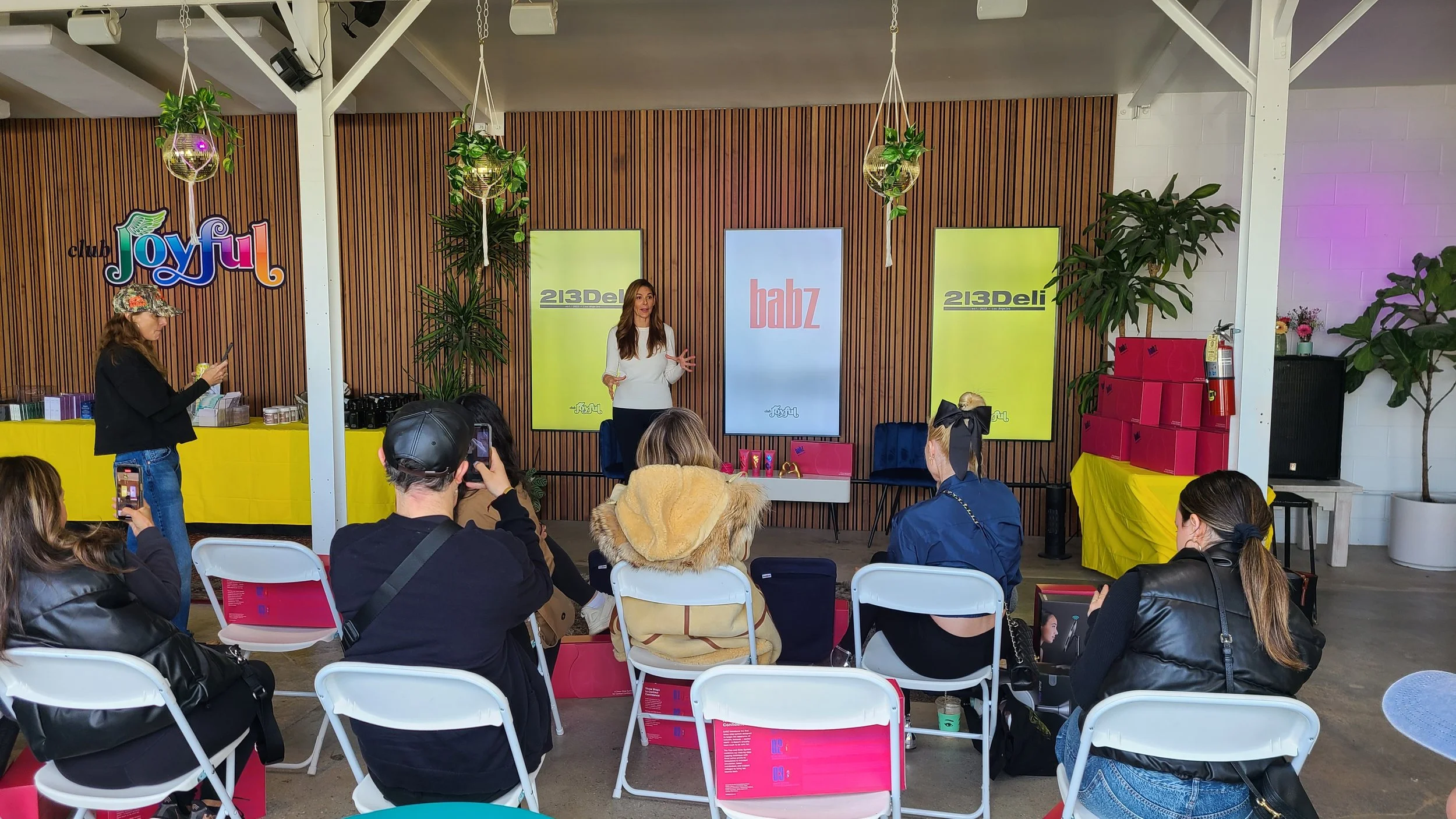 A woman is standing on a stage, giving a presentation to an audience seated in chairs. The backdrop includes colorful banners with the words 'Babz,' '213Del,' and 'Joyful.' There are plants and a display table on either side of the stage, with some b