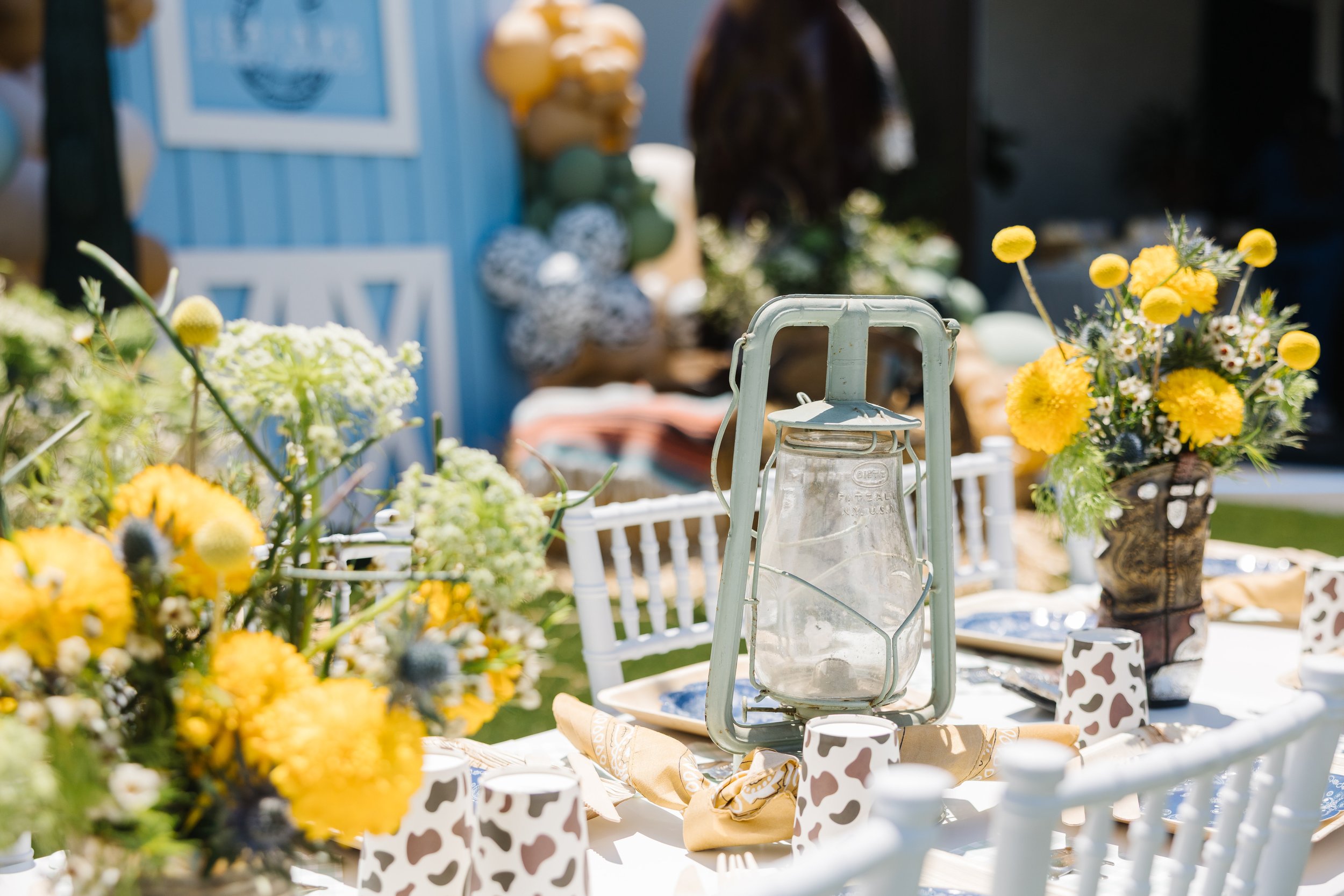 Outdoor table setting with yellow and white floral arrangements, a vintage lantern, and cow print cups.