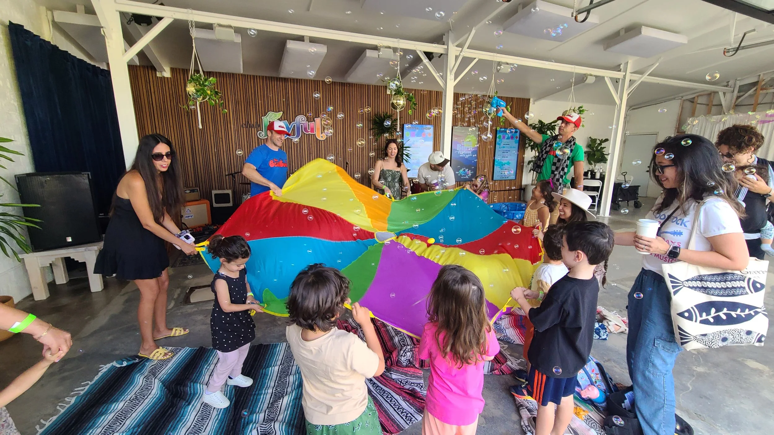 Children and adults participating in a parachute game during a party, with bubbles floating in the air and a party room setting.