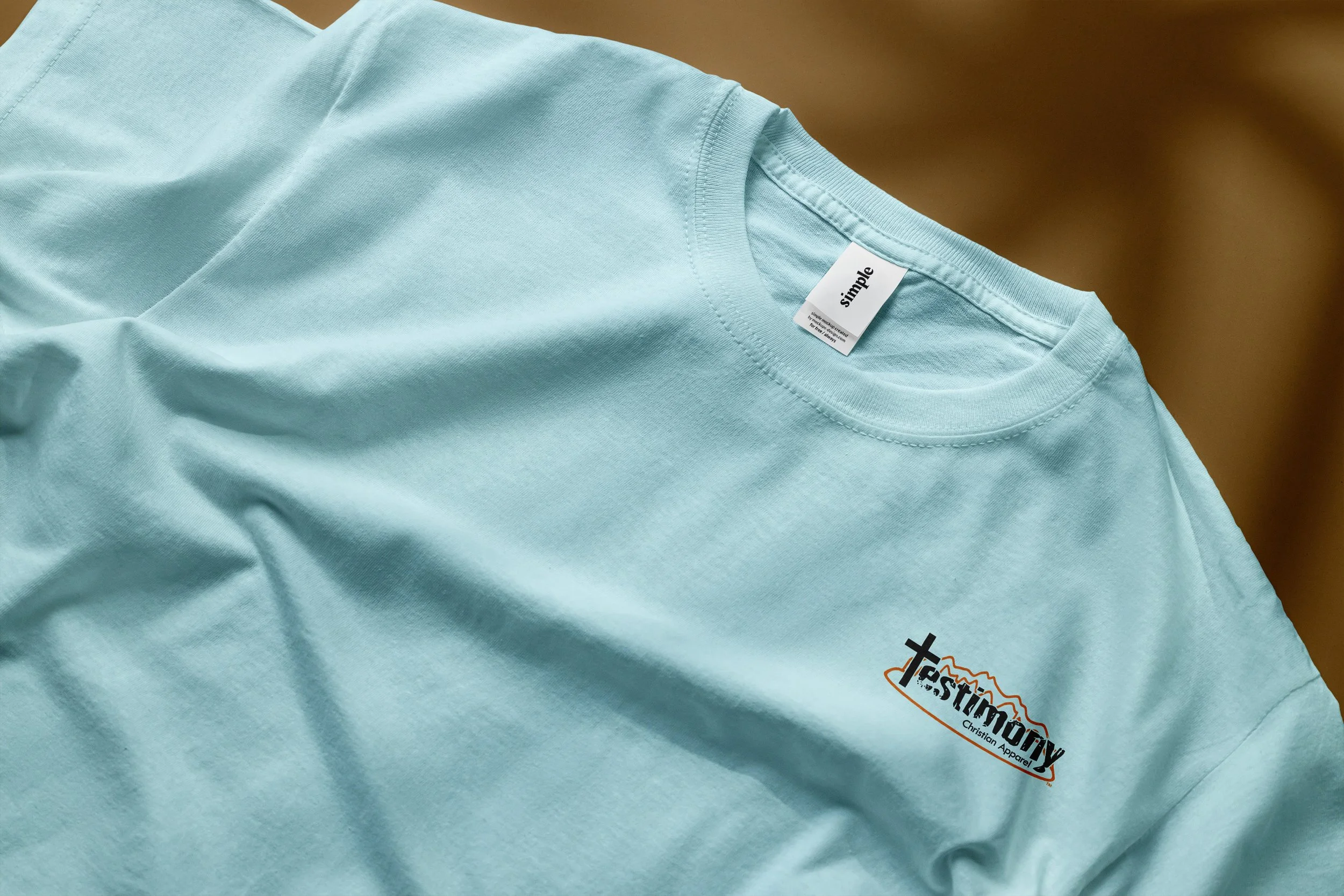 Close-up photo of t-shirt laid flat showing Testimony Christian Apparel logo on front left chest