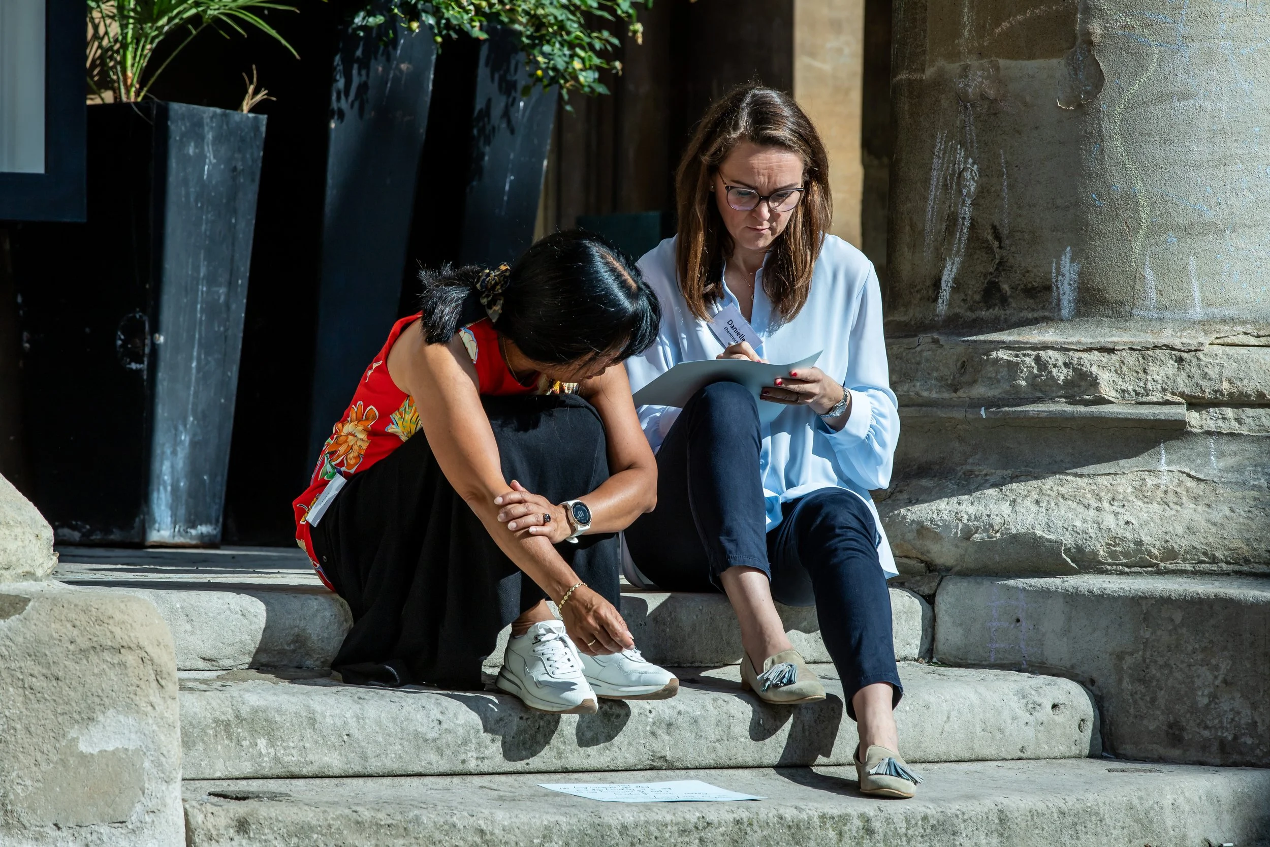 Two women sitting on stone steps outdoors, one helping the other with her sneaker.