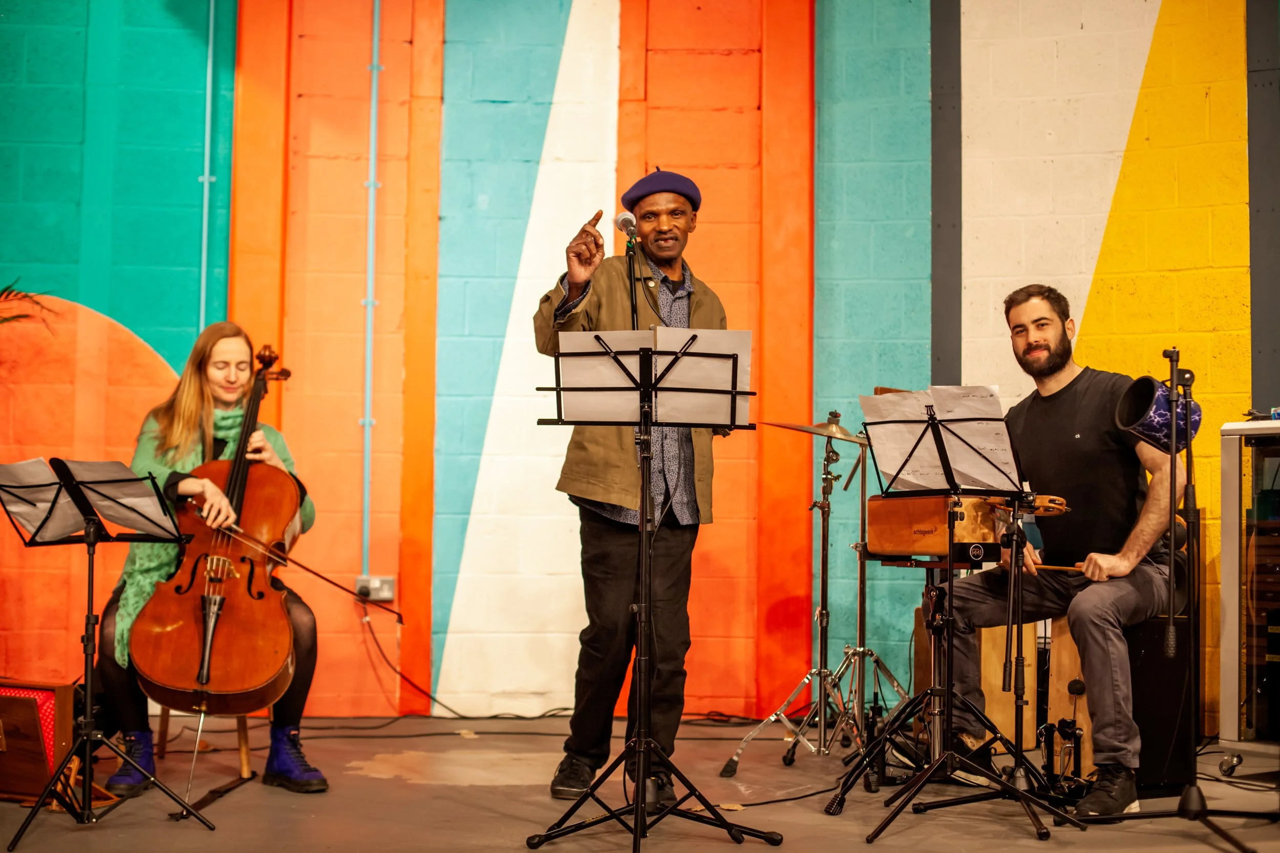 Three musicians performing on a stage with a colorful striped background. The woman on the left is playing a cello, the man in the middle is speaking into a microphone, and the man on the right is playing percussion with a cajón.