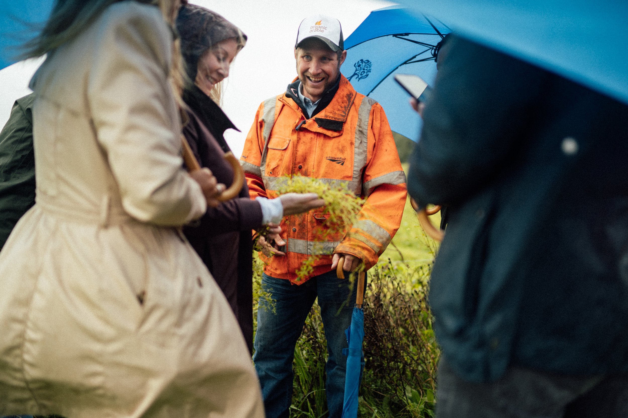 A group of people outdoors in rainy weather, with one man in an orange rain jacket smiling, holding a small plant, and others holding umbrellas.