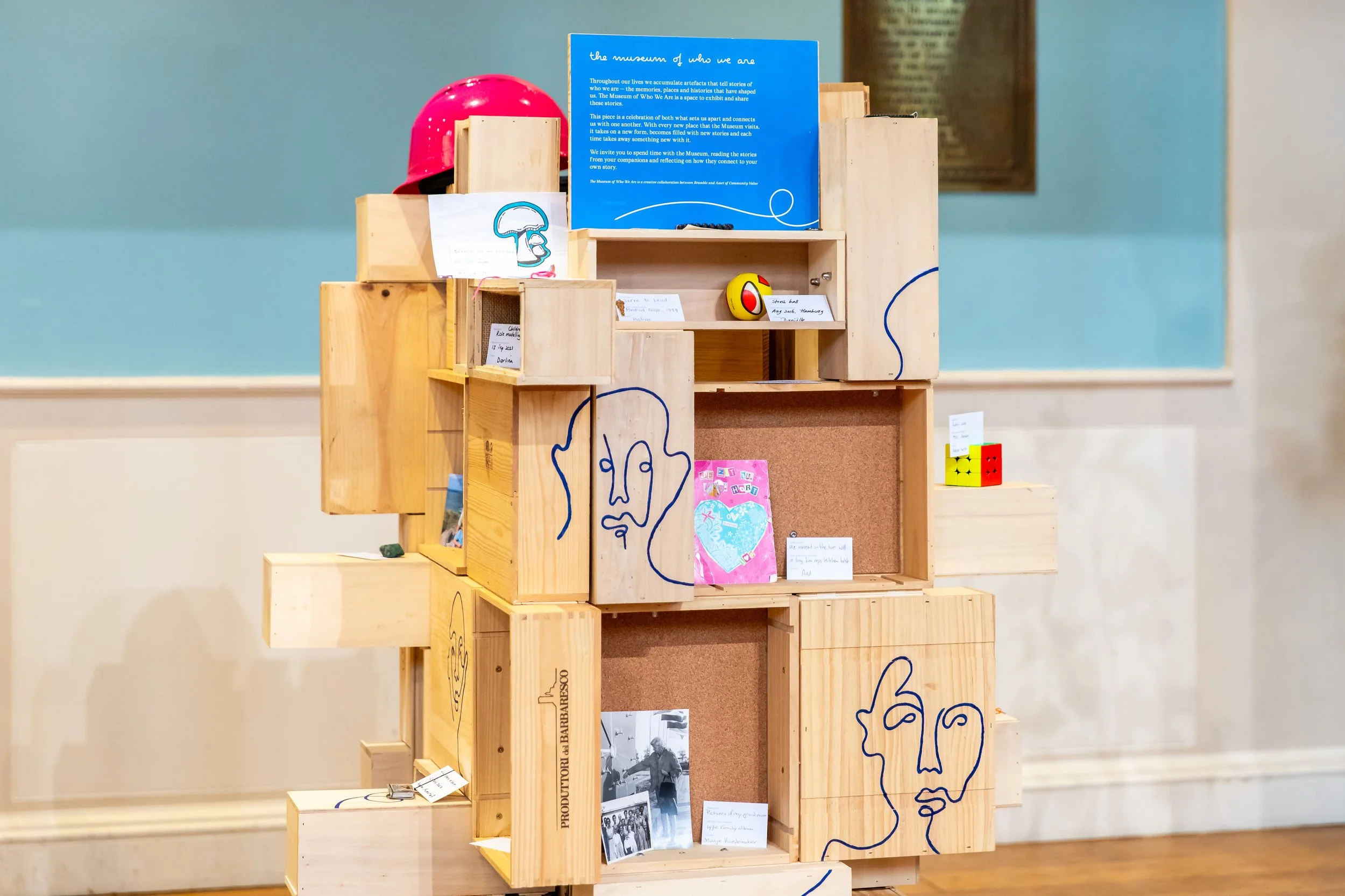 A wooden sculpture with various compartments, art, and small objects on display, decorated with line drawings of faces.