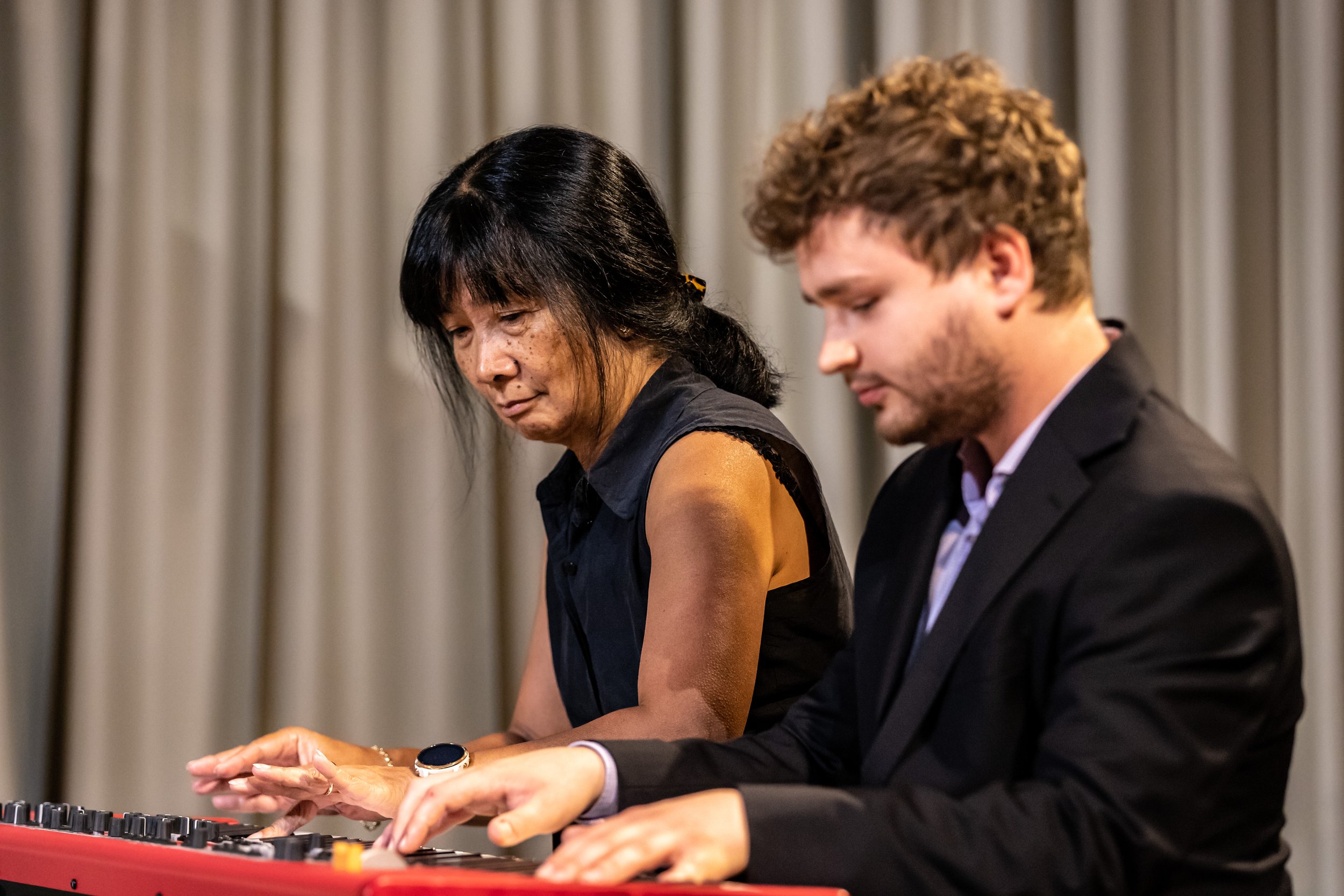 A woman and a man playing a red keyboard together, with a beige curtain in the background.