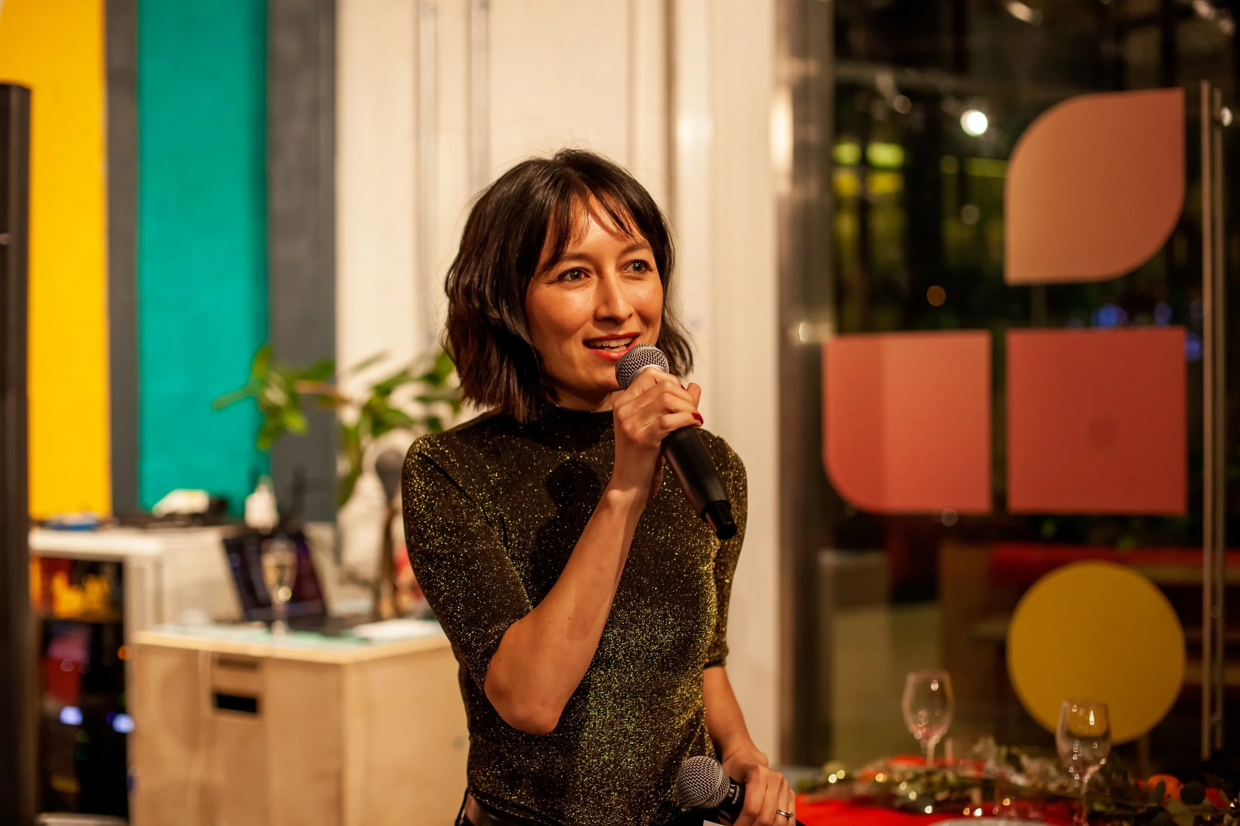 A woman with shoulder-length dark hair holding a microphone and speaking in a room decorated with colorful shapes and a table with glasses and bottles.