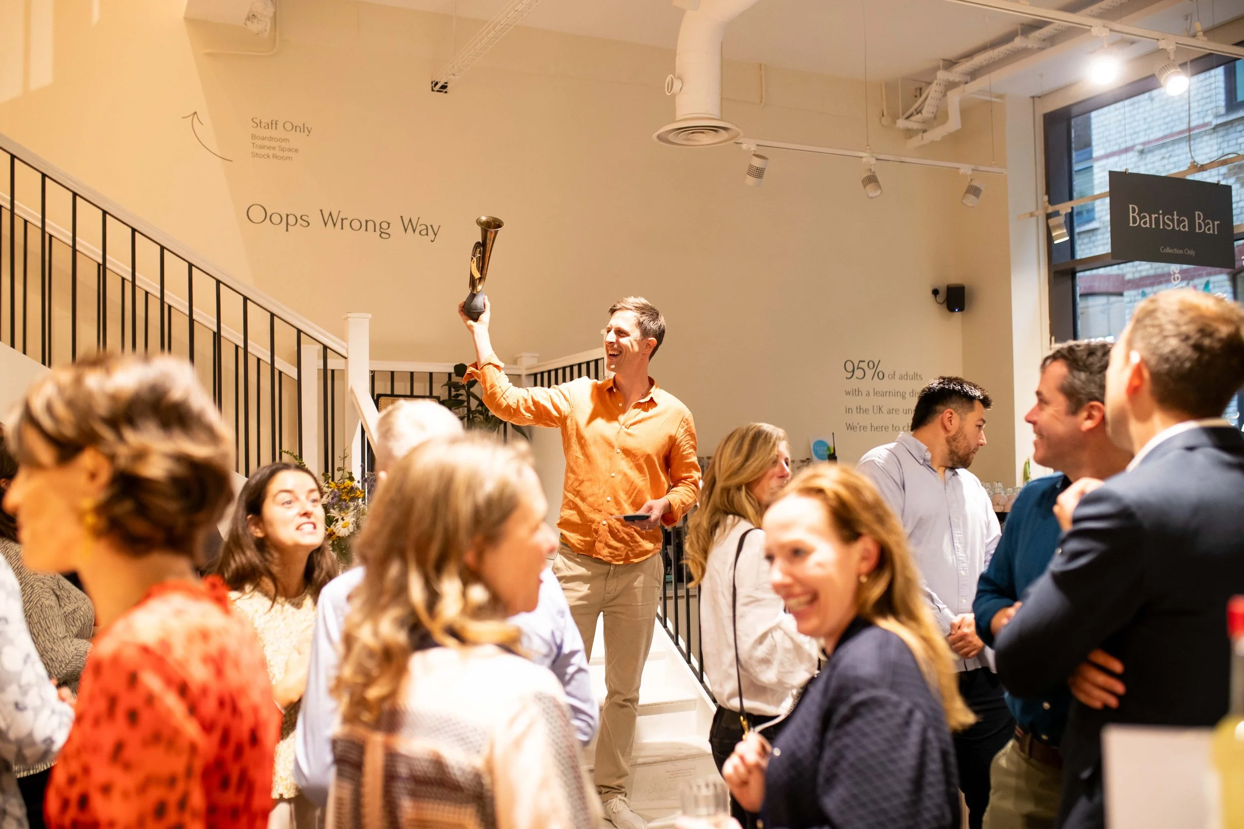A man in an orange shirt holding a trophy in the air while smiling at a social gathering in a modern indoor space with several people engaged in conversation and smiling.
