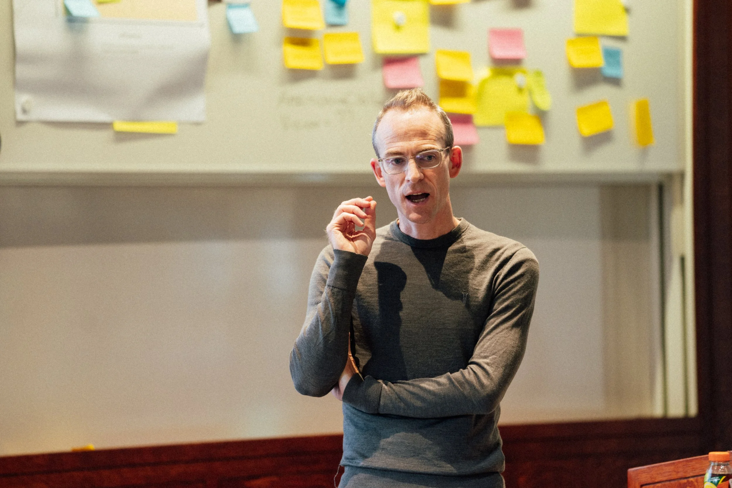 A man with glasses and short hair is talking or giving a presentation in front of a wall covered with colorful sticky notes and paper sheets.