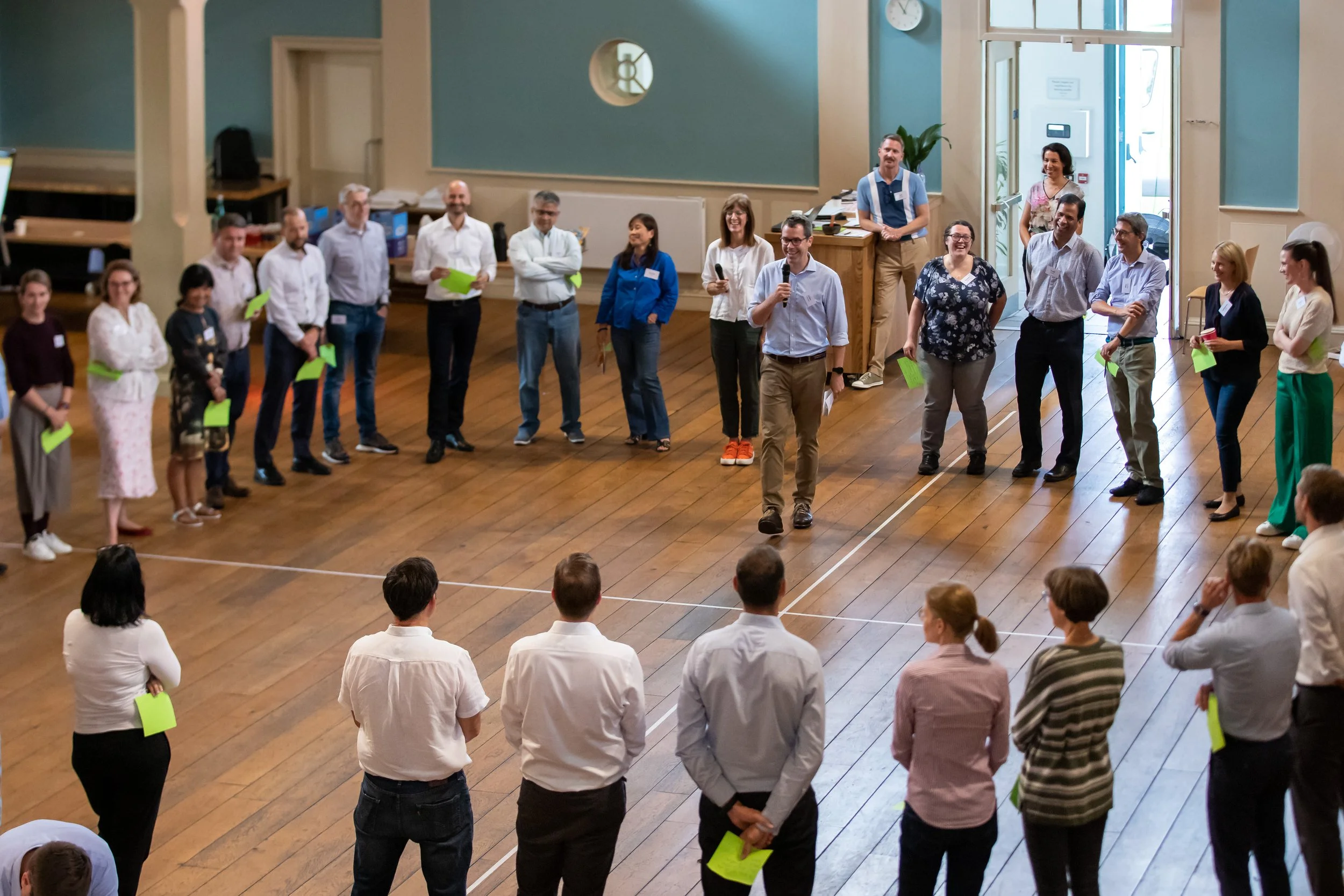 Group of people participating in a team-building activity indoors, forming a large circle, with a man in the center holding a microphone.