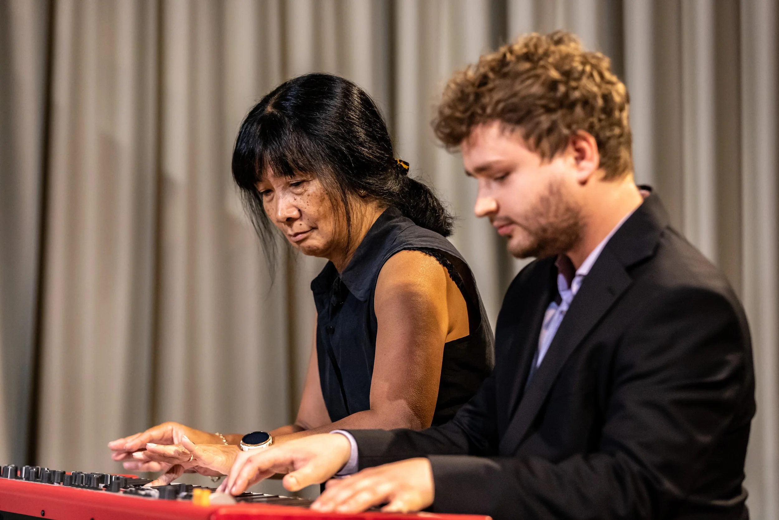A woman and a man playing a red keyboard together, with a beige curtain in the background.