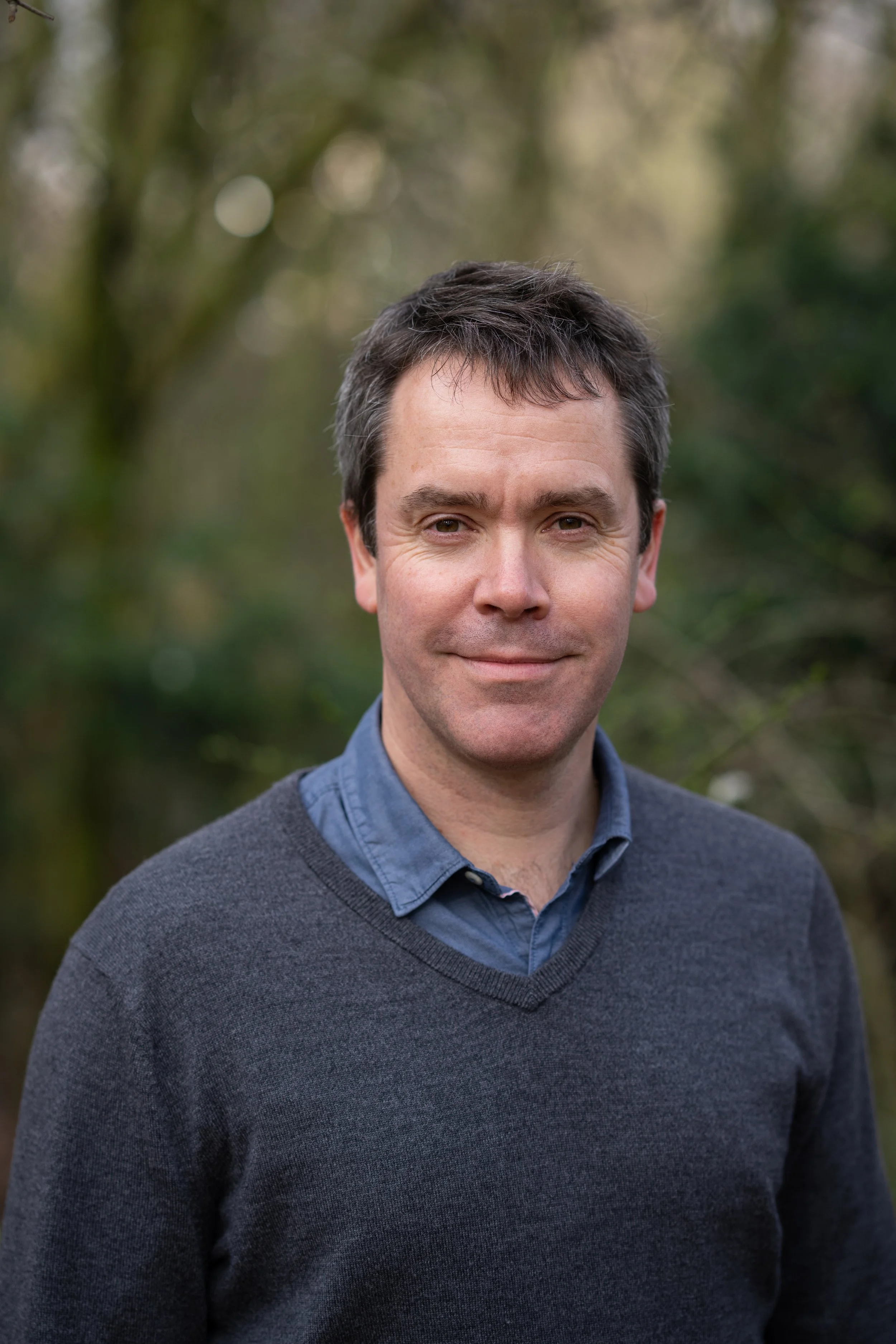 A man outdoors in a natural setting with blurred trees in the background, wearing a blue collared shirt and a dark sweater, smiling softly.