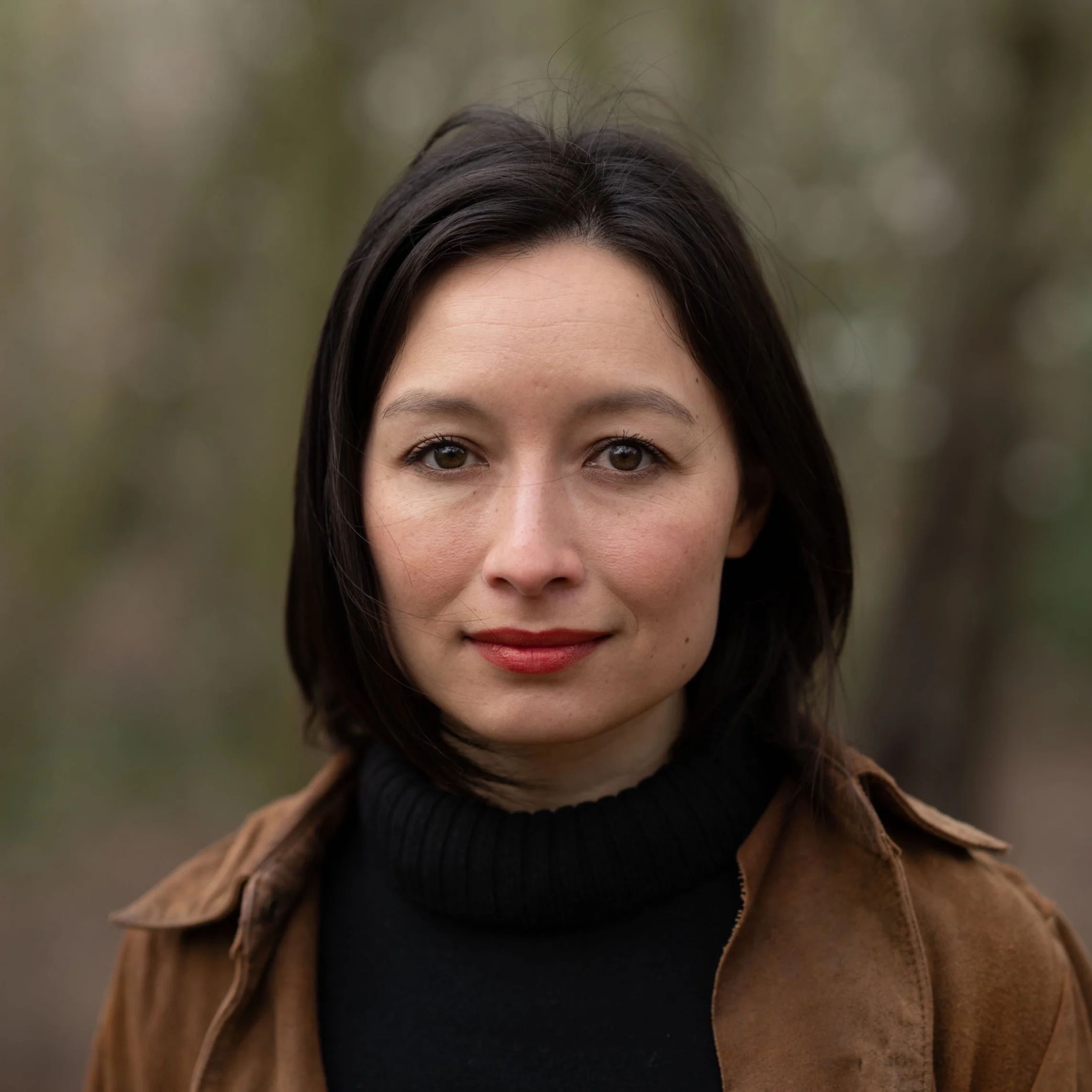 Portrait of a woman with dark hair and brown eyes, wearing a black turtleneck and brown jacket, outdoors with blurred trees in the background.
