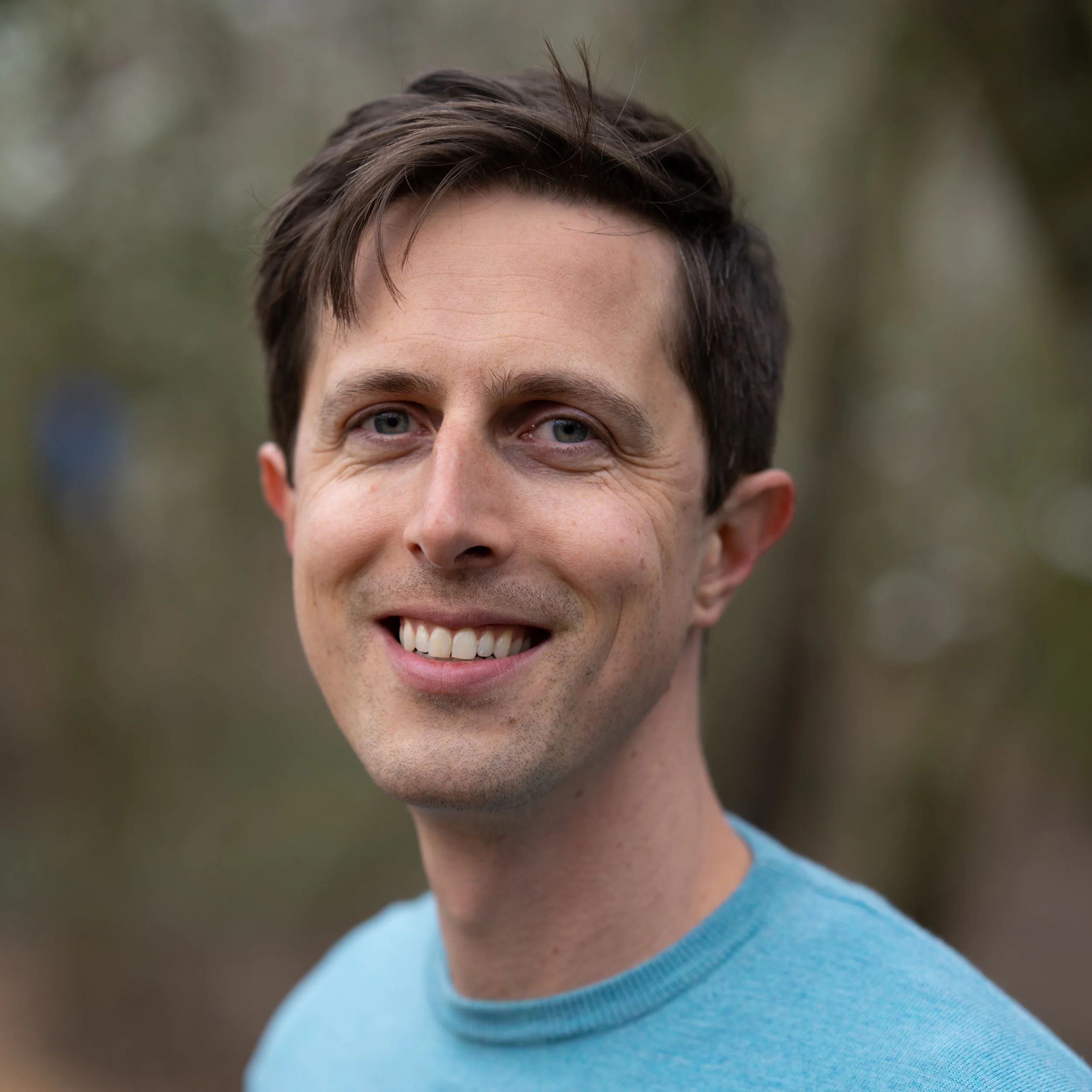 Close-up of a smiling man with dark brown hair, blue eyes, and fair skin, wearing a light blue shirt, outdoor background with blurred greenery.