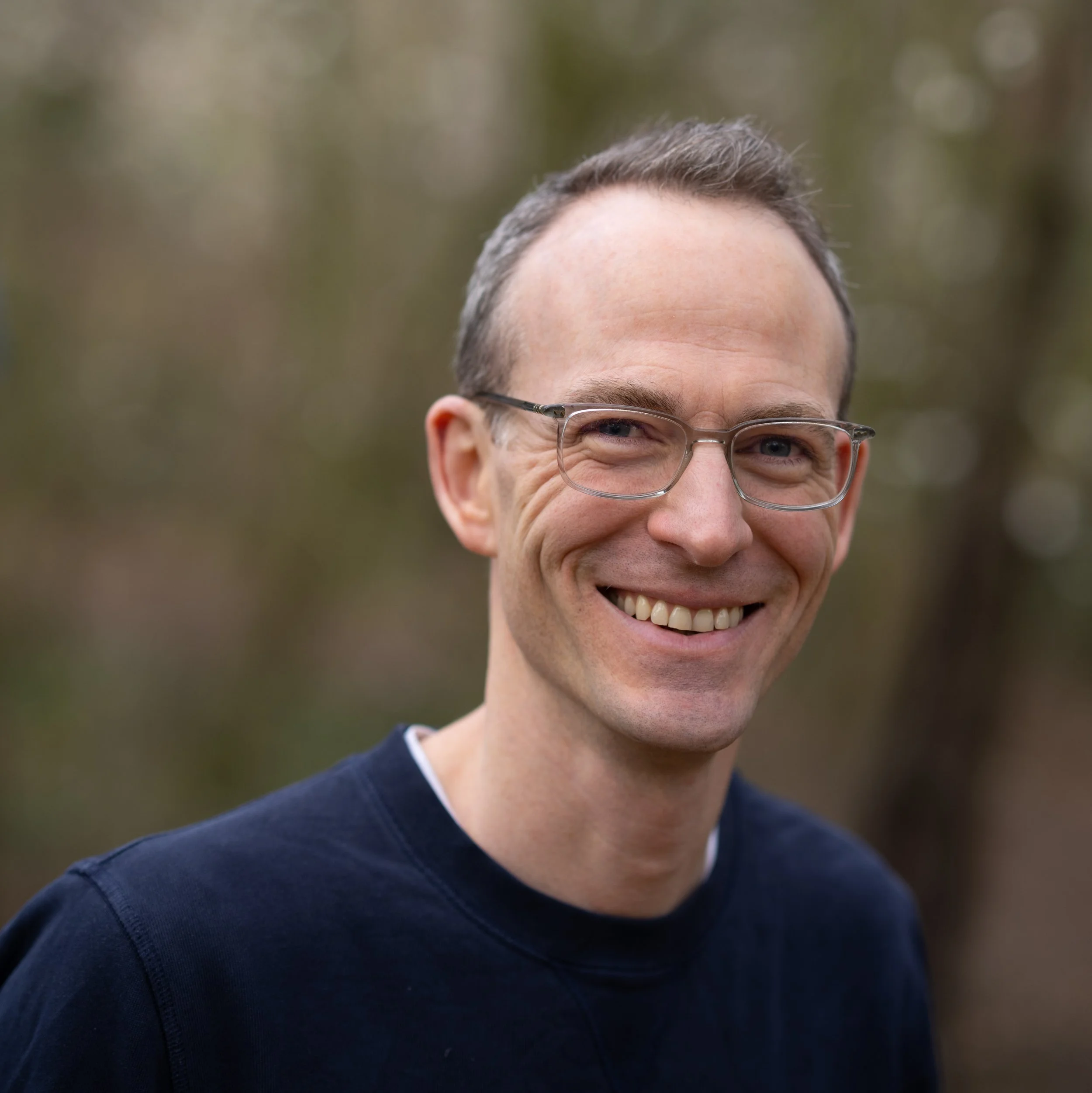 A smiling man with glasses and short hair outdoors, wearing a dark shirt.