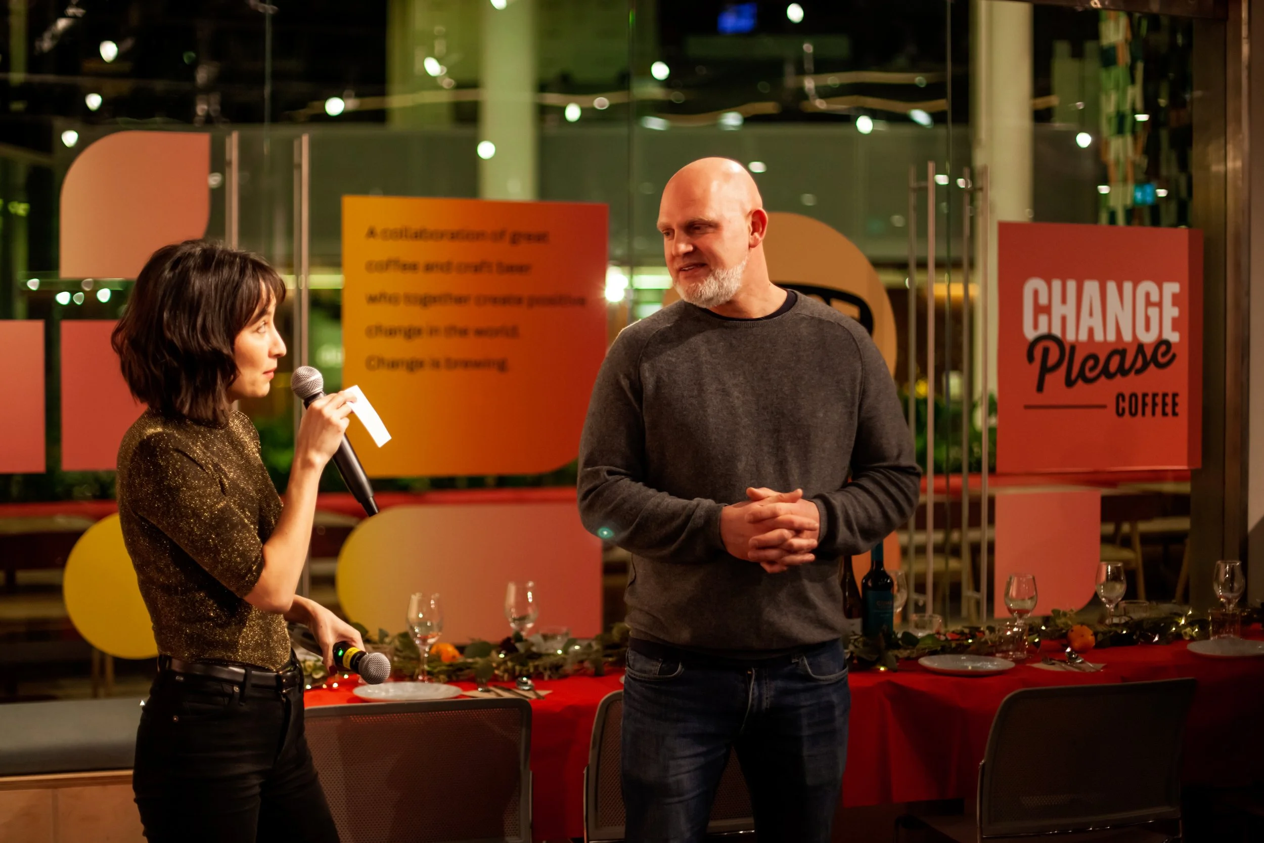 A woman holding a microphone and speaking to a man with a beard, indoors at a decorated event with signs that say 'Change Please Coffee' in the background.