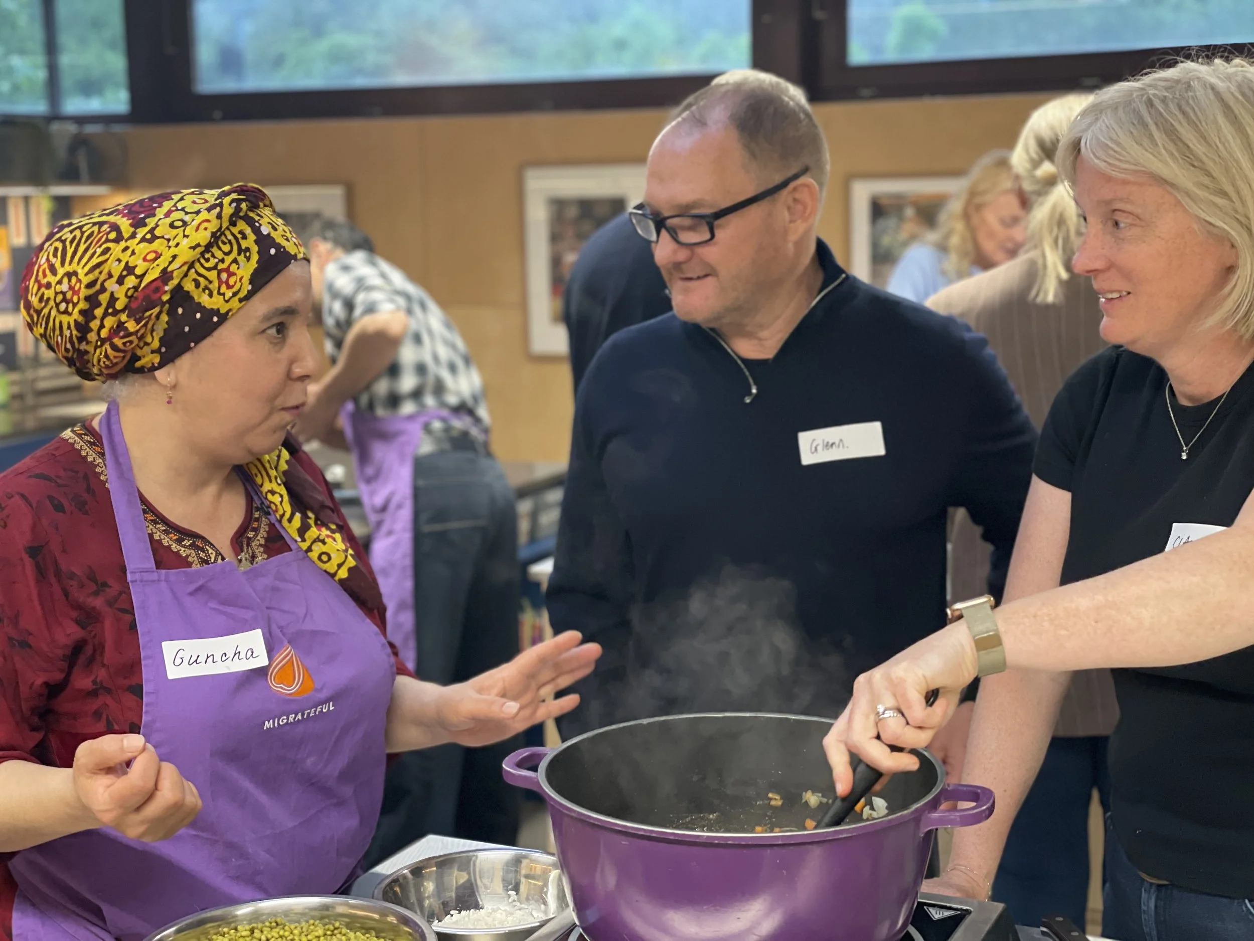 Three people engaging in a cooking demonstration: a woman wearing a colorful headscarf and purple apron labeled 'Guncha,' a man with glasses and a black shirt labeled 'Glen,' and a woman in a black shirt, stirring food in a purple pot.