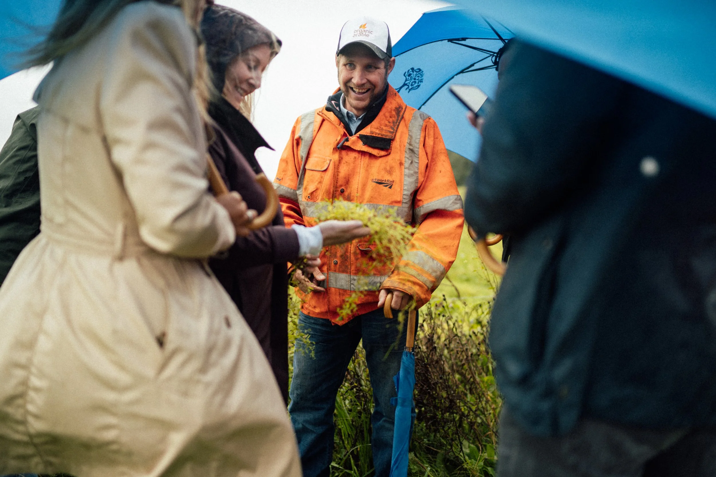 A group of people outdoors, some holding umbrellas in the rain, standing on grass. They are gathered around a man in an orange safety jacket, who is smiling and holding a small plant or sprig.