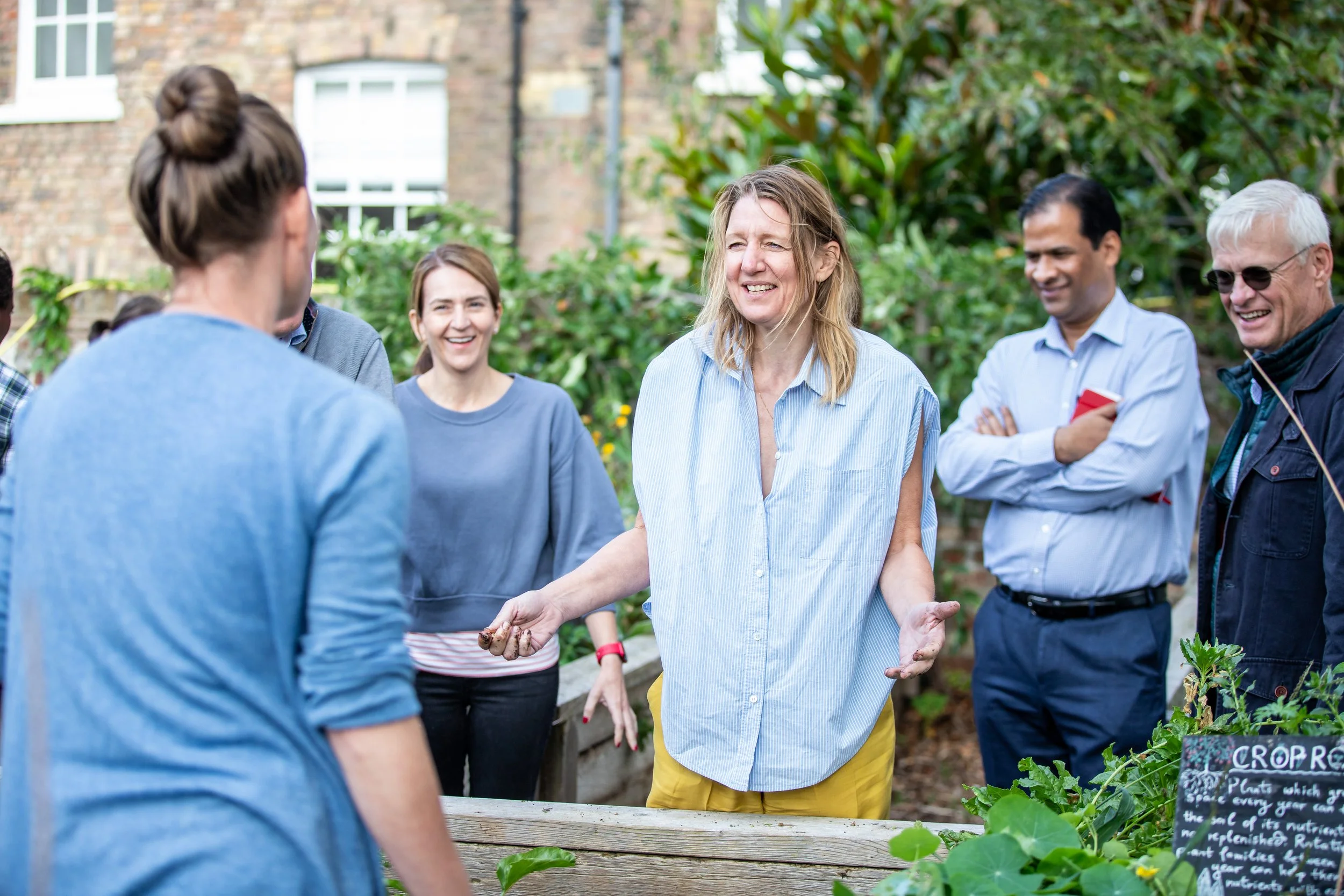 Group of people gathered outdoors in a garden, smiling and engaging in conversation, with a woman in the center talking.