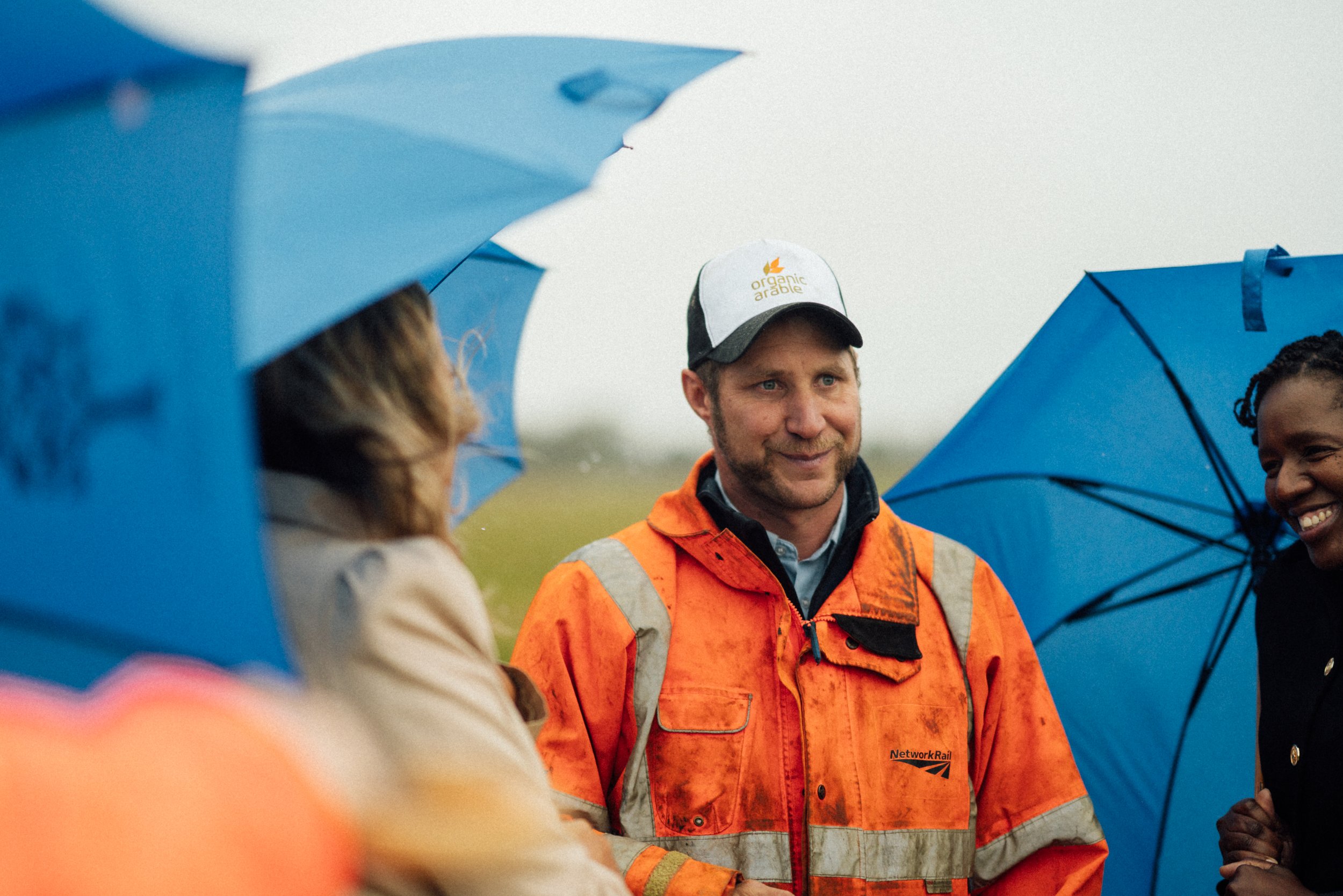 People gathered outdoors holding blue umbrellas, with a man in the center wearing an orange high-visibility jacket and a white cap, engaging in conversation.