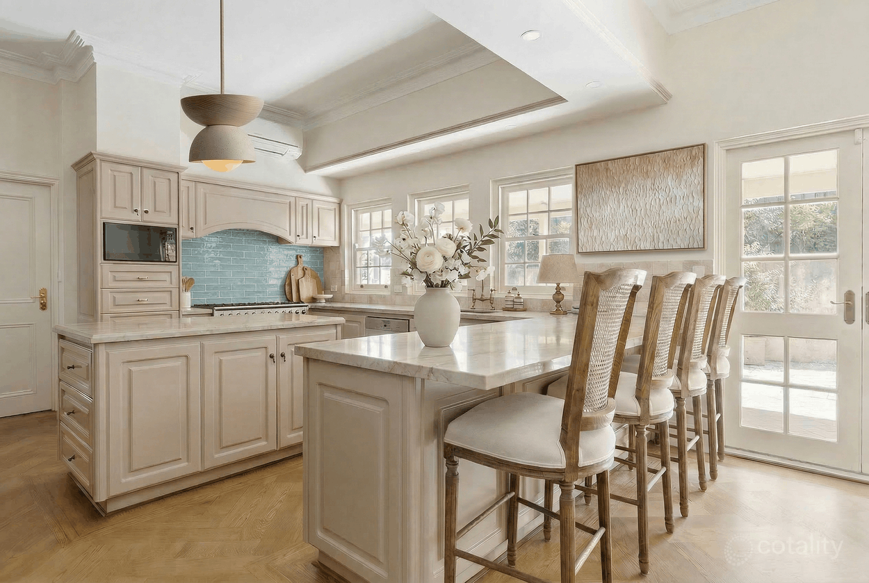 Bright kitchen with white cabinetry, marble island, blue tiled backsplash, and a row of beige chairs at the island. There is a vase with white flowers on the island and large windows letting in natural light.