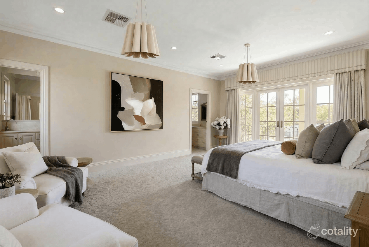 A bedroom with a large bed covered in white bedding and multiple pillows, near a window with curtains, a small sofa with a dark throw, a side table with a floral arrangement, and abstract artwork on the wall.