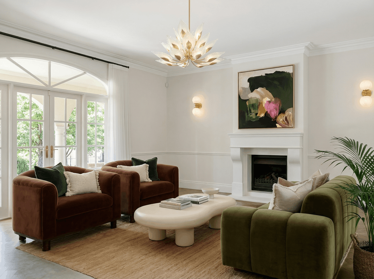Living room with white walls and ceiling, four velvet sofas in green and brown around a white coffee table, large window with white curtains, abstract painting above a white fireplace, decorative pendant light, and indoor plant.