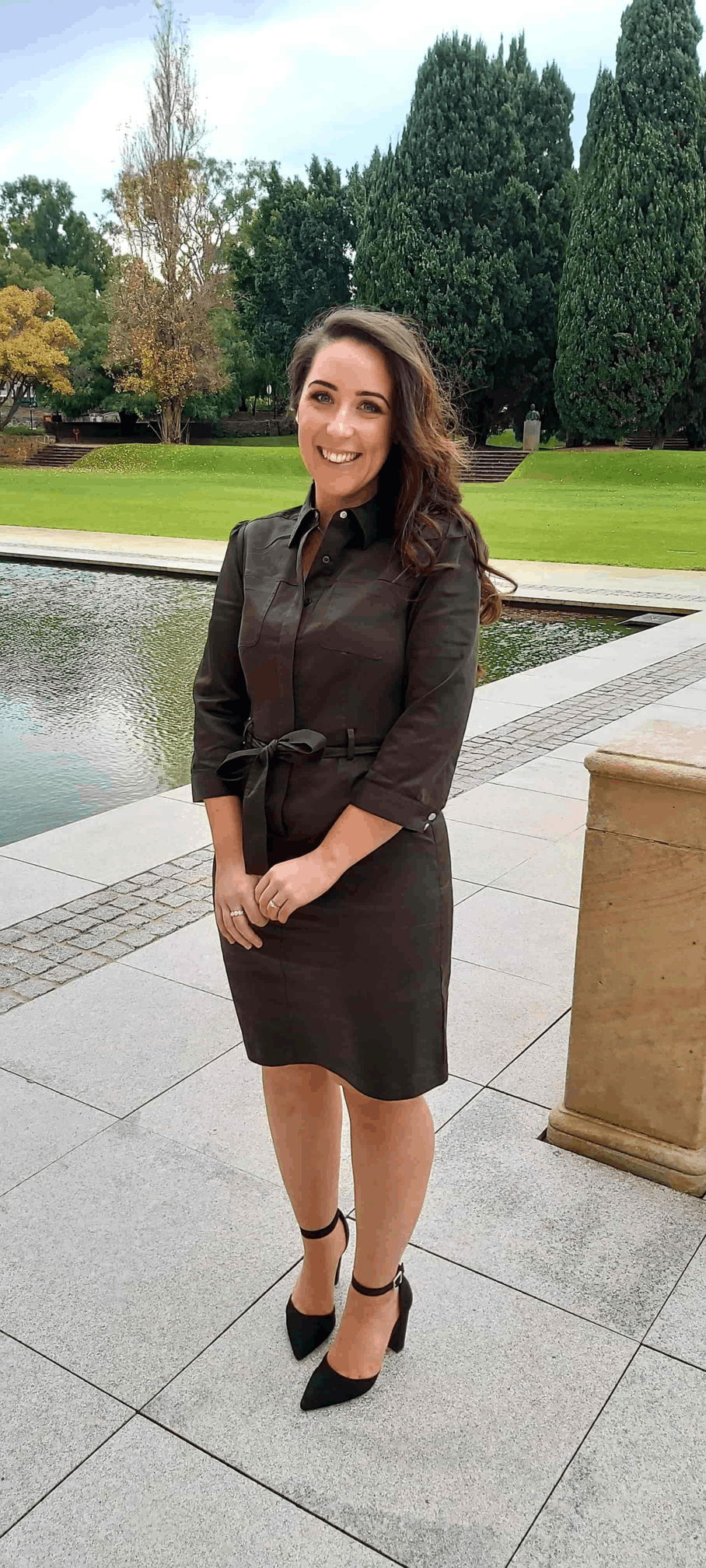 Jodie Townsend, interior designer, standing outdoors by a tranquil pond surrounded by lush green trees, wearing a black dress and high heels.