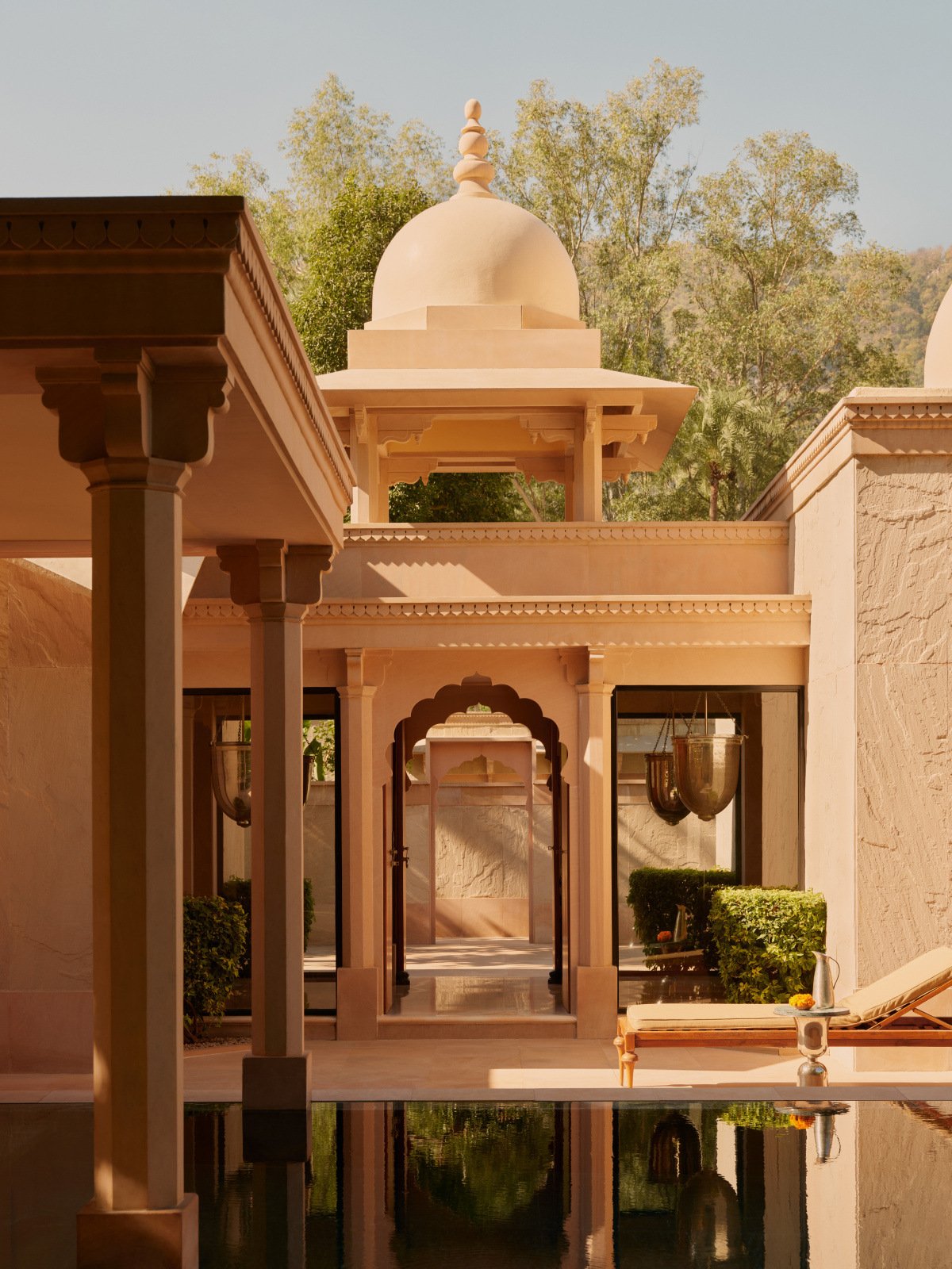A view of an Indian-style building with light pink walls, domed roof, and arches. There are hanging lamps, plants, and a swimming pool, with trees in the background. Discover India with Aman at Amanbagh Rajastan, and Aman-i-Khás Ranthambore National.