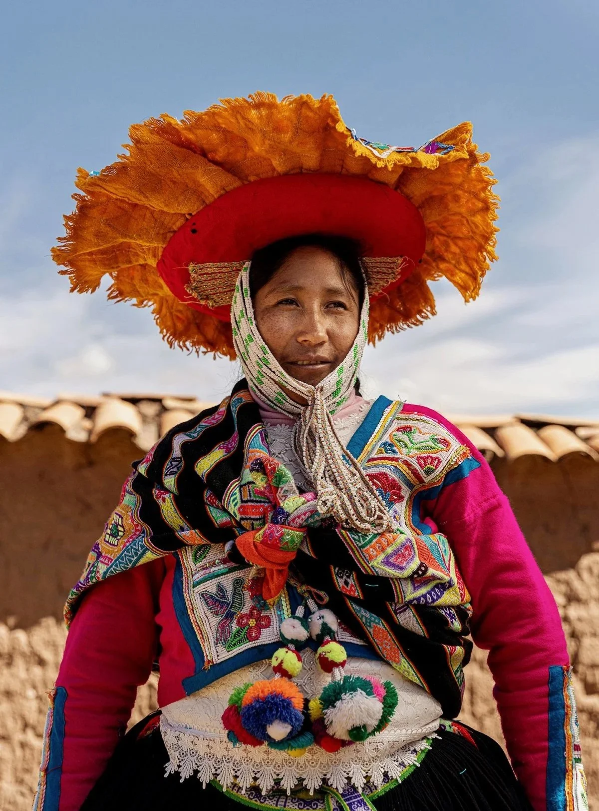 A woman wearing traditional, colorful Andean clothing and a large, orange, and red hat with fringe, standing outdoors against a blue sky and a mud-brick wall. Traditional, culture, adventure travel in Peru, Atacama, Chile, Patagonia.