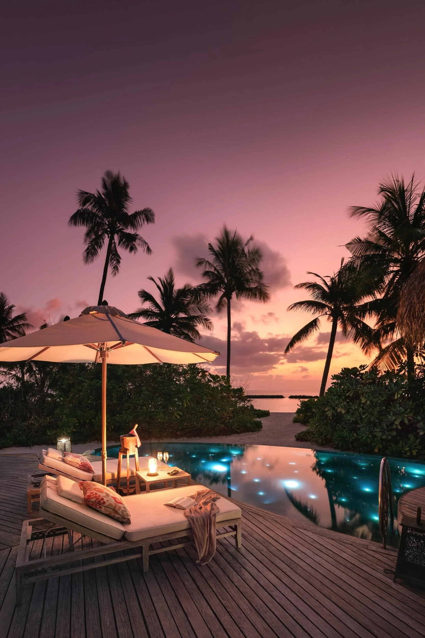 Romantic tropical resort scene during sunset with palm trees, a pool with underwater lighting, lounge chairs with pillows and blankets, a large white umbrella, and small tables with candles and drinks. Romantic travel at The Nautilus, Maldives.