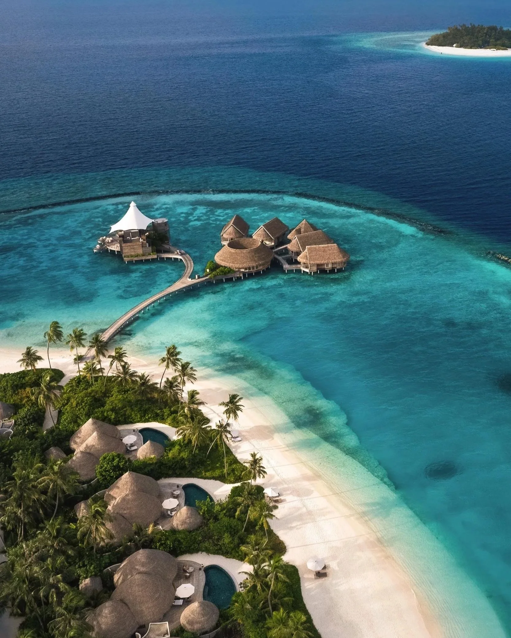 An aerial view of overwater bungalows with thatched roofs on a tropical island, surrounded by clear turquoise water and a sandy beach with palm trees. Located at The Nautilus, Maldives.