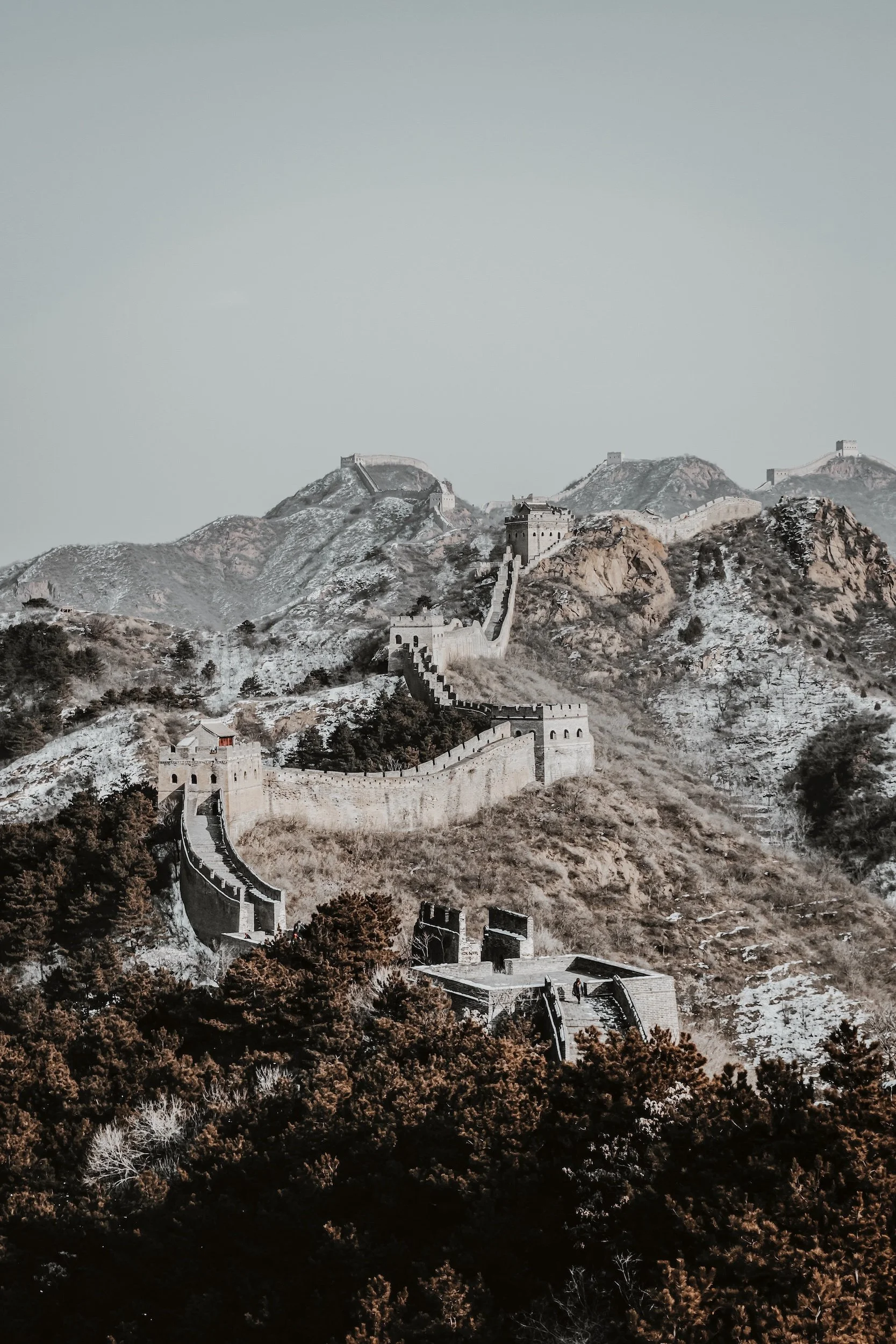 Snow-covered section of the Great Wall of China winding through rocky, foggy mountains with a pale, overcast sky. Traditiona, historical and culturearcheological travel adventures in Africa, Egypt, Asia and Machu Pitchu.