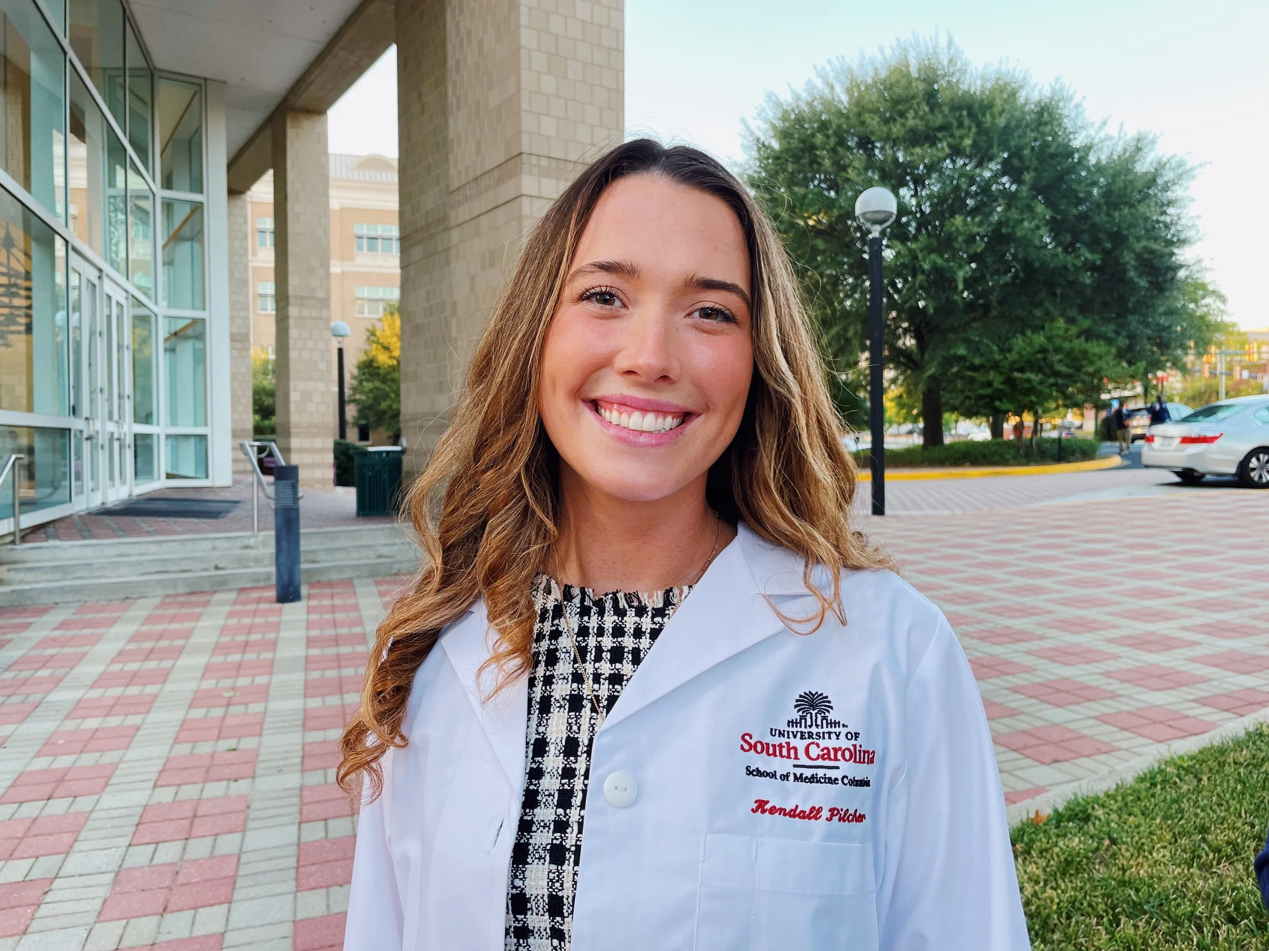 A young woman in a white lab coat standing outdoors on a university campus, smiling, with trees, a sidewalk, and cars in the background.