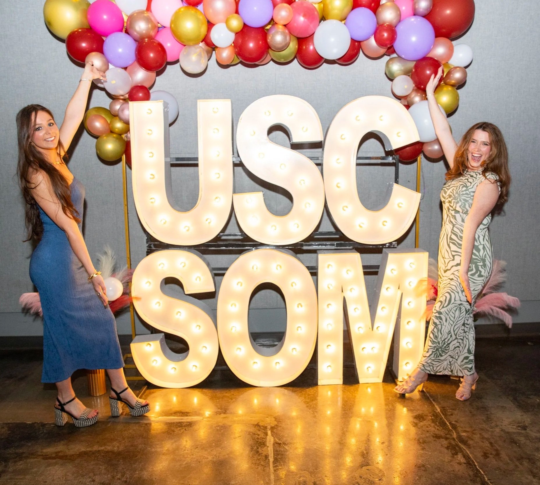 Two women dressed in stylish dresses standing beside a large illuminated sign that reads "USC SOM" with colorful balloons arranged above and around the sign.