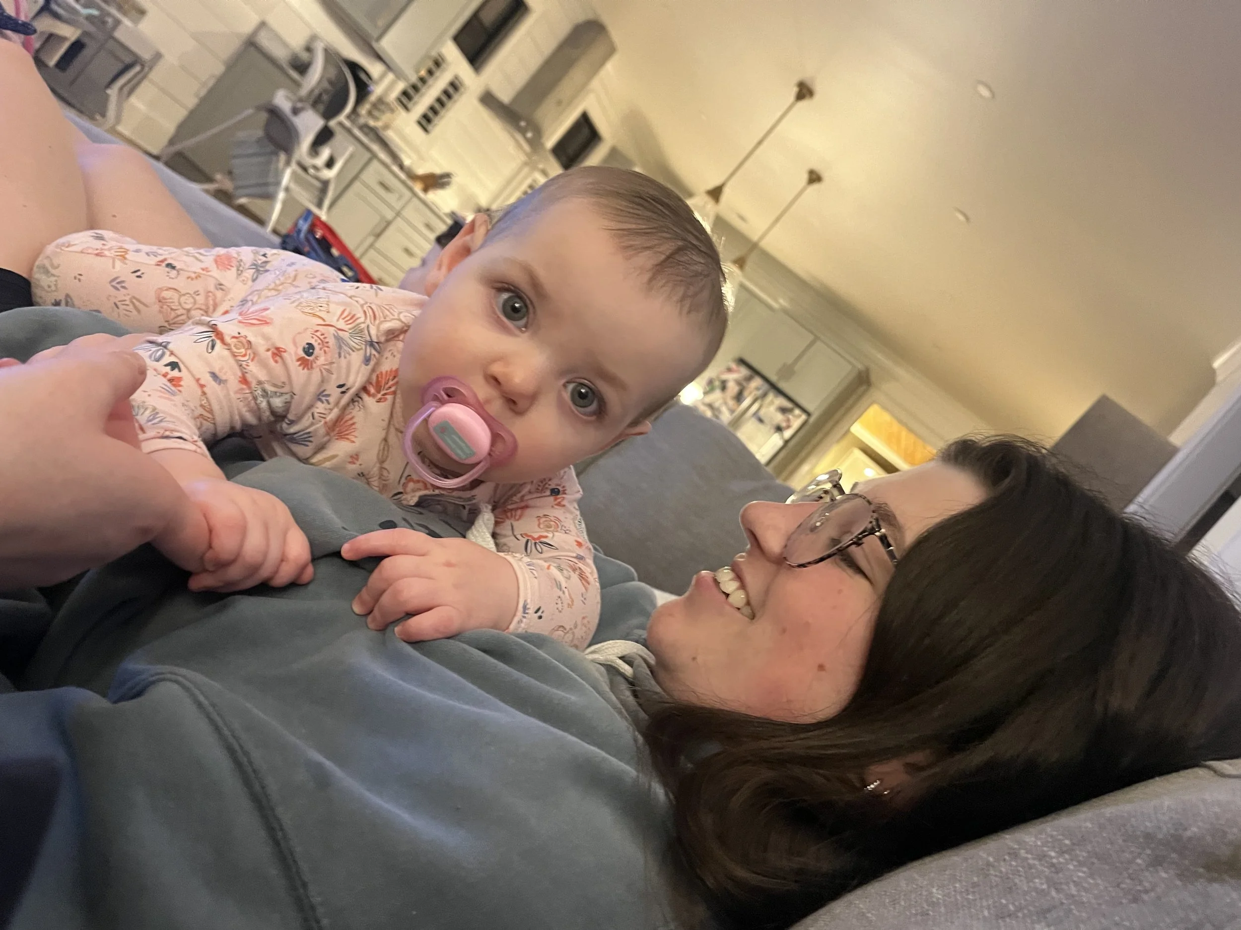 A young child with blue eyes and a pacifier in their mouth lying on top of a woman, likely their mother, who is smiling and wearing glasses. They are in a living room with a kitchen in the background.