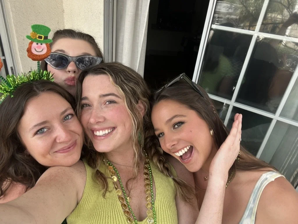Four young women celebrating St. Patrick's Day, wearing festive accessories like green necklaces and leprechaun hats, smiling and taking a selfie outside near a glass door.