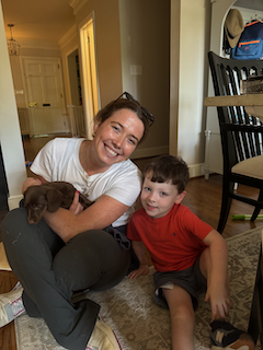 A woman sitting on the floor holding a small brown puppy, with a young boy in a red shirt sitting beside her, in a home interior with chairs and a doorway in the background.