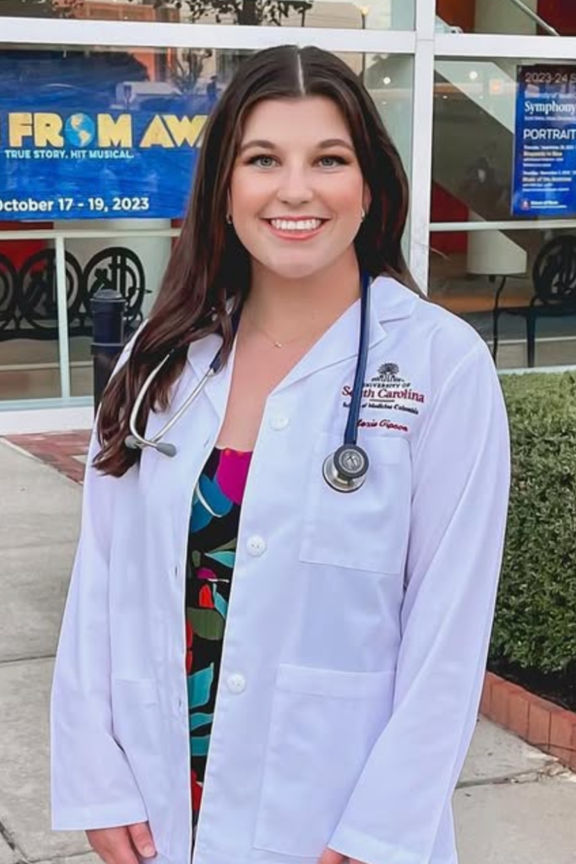 A young woman in a white doctor's coat with a stethoscope around her neck, smiling outside a building with posters in the background.