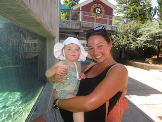 A woman holding a young girl, both wearing summer clothes, near a swimming pool with trees and a building in the background.