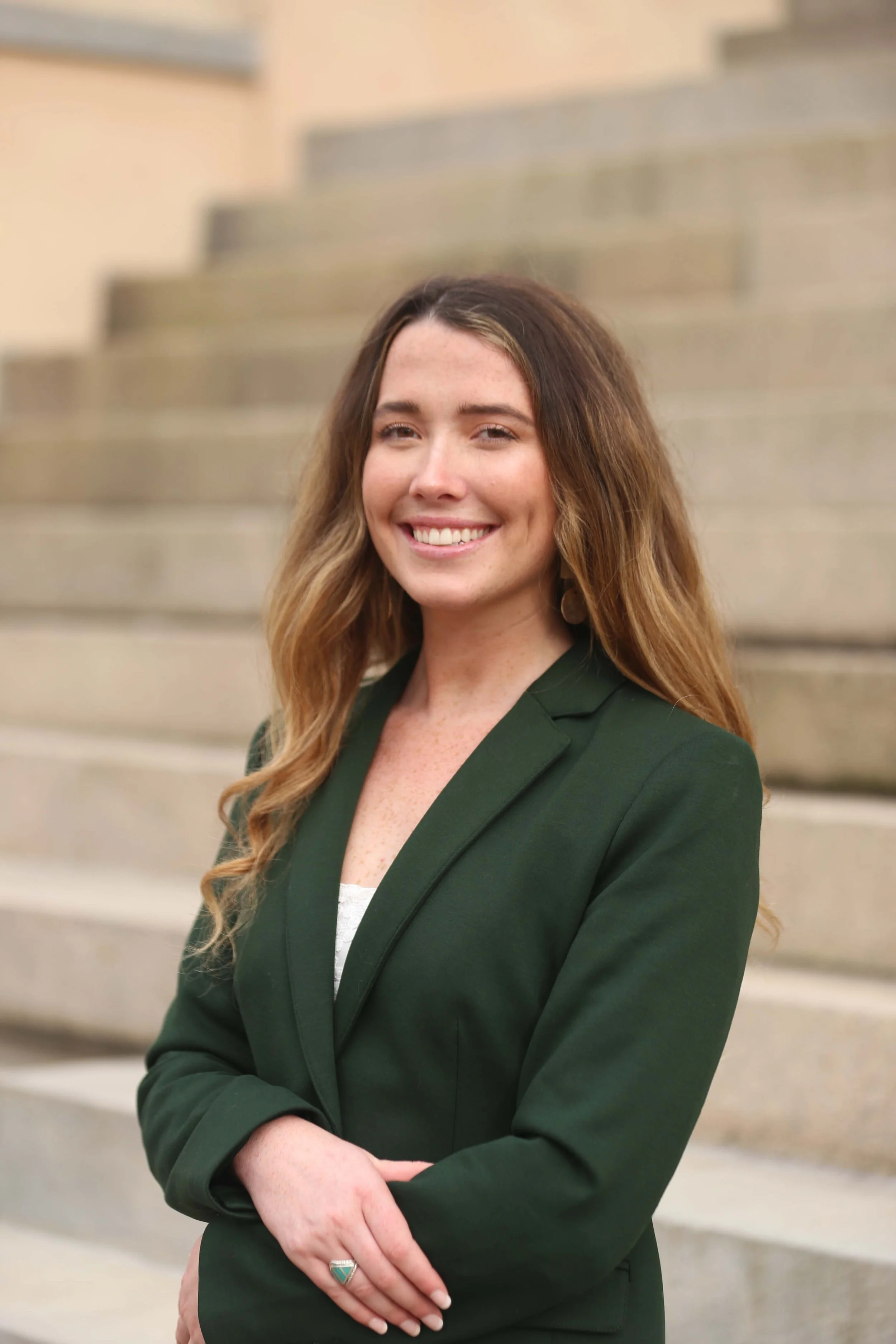 A woman with long wavy brown hair, smiling, standing outdoors in front of stone stairs, wearing a dark green blazer and a ring on her left hand.