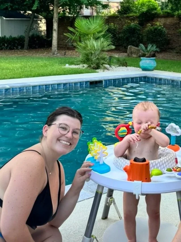 A woman and a young child in a baby float in a backyard swimming pool. The woman is smiling and wearing glasses and a black swimsuit. The child is playing with toys, holding a yellow rattle, and has a little floaty on around the waist. The pool has a blue tile border, and there are trees, bushes, a fenced yard, and a decorative plant in a blue pot in the background.