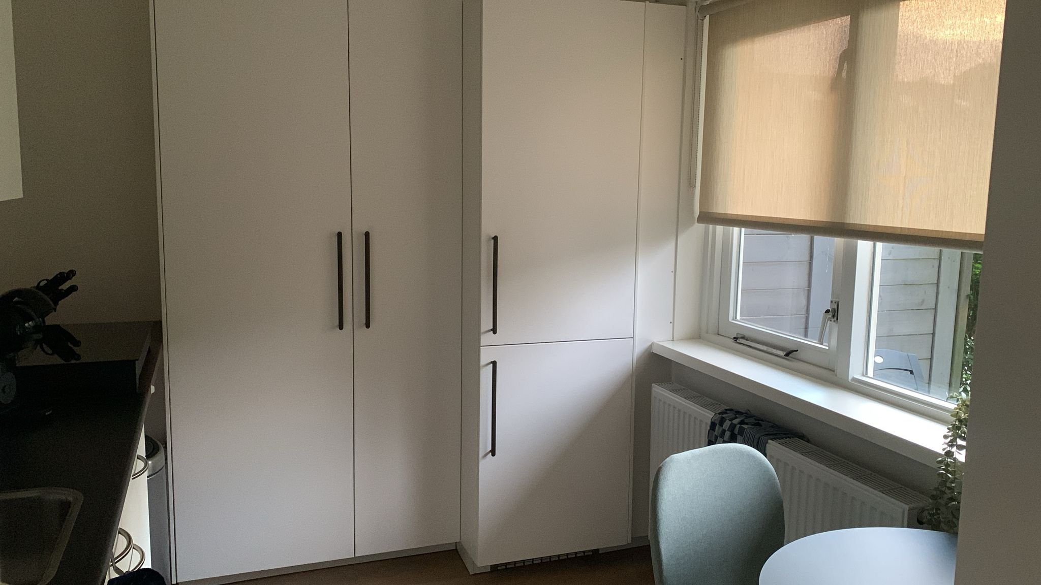 Een keukenhoek met witte kasten, een raam met een beige rolluik en een stoel naast een ronde tafel.