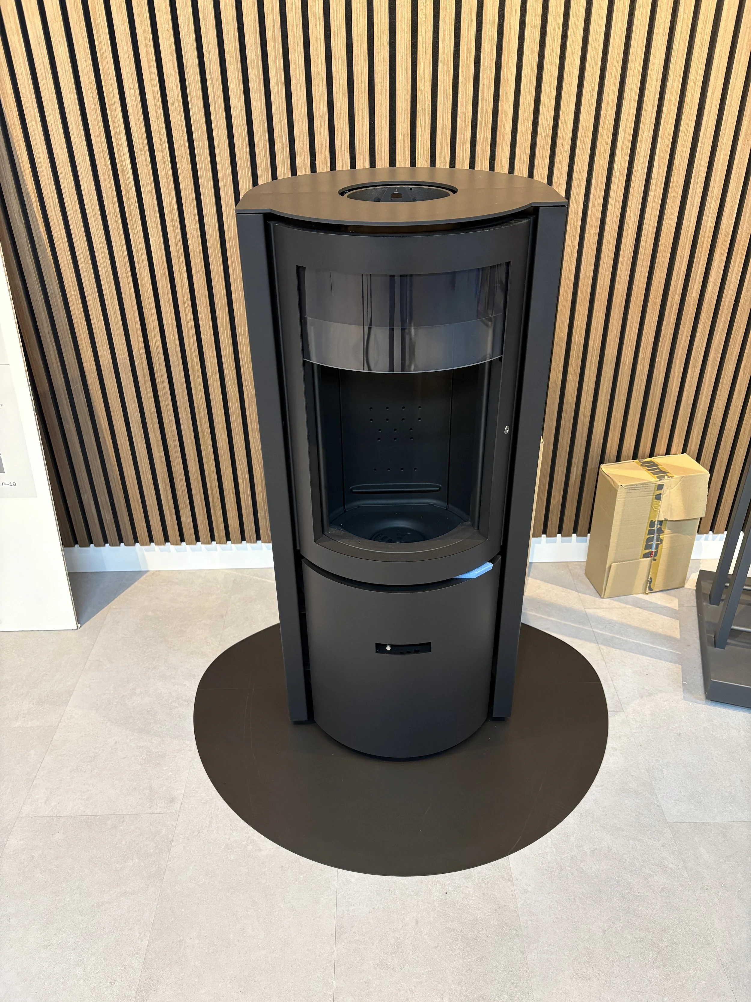 een zwart ronde toestel in een winkel of showruimte, geplaatst op een vloer met een donkere cirkel eronder, tegen een houten wand met verticale strepen.