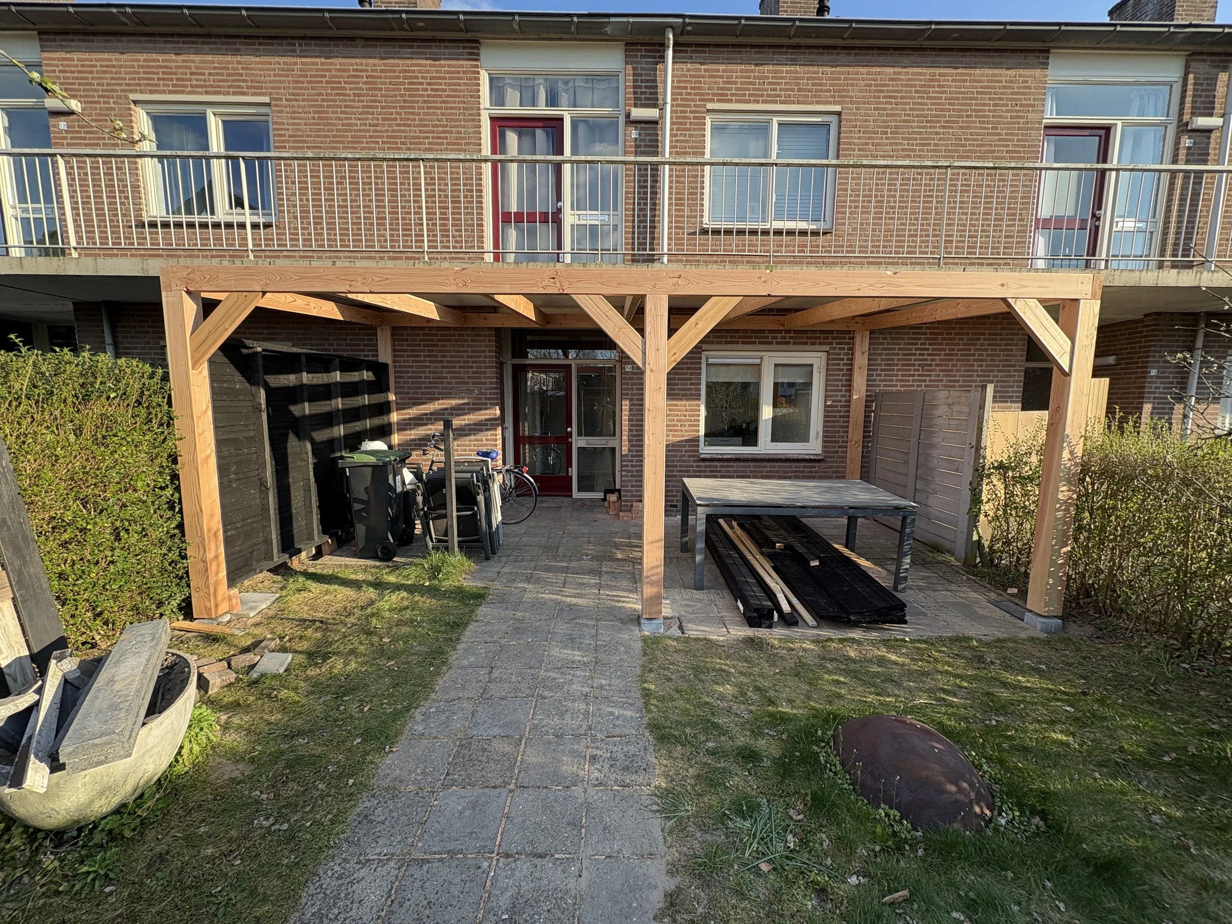 Bijkomende aanbouw in aanbouw met houten frame onder constructie. Tuin met bestrating en planten. Achterzijde van een appartementengebouw met balkondeuren en ramen.